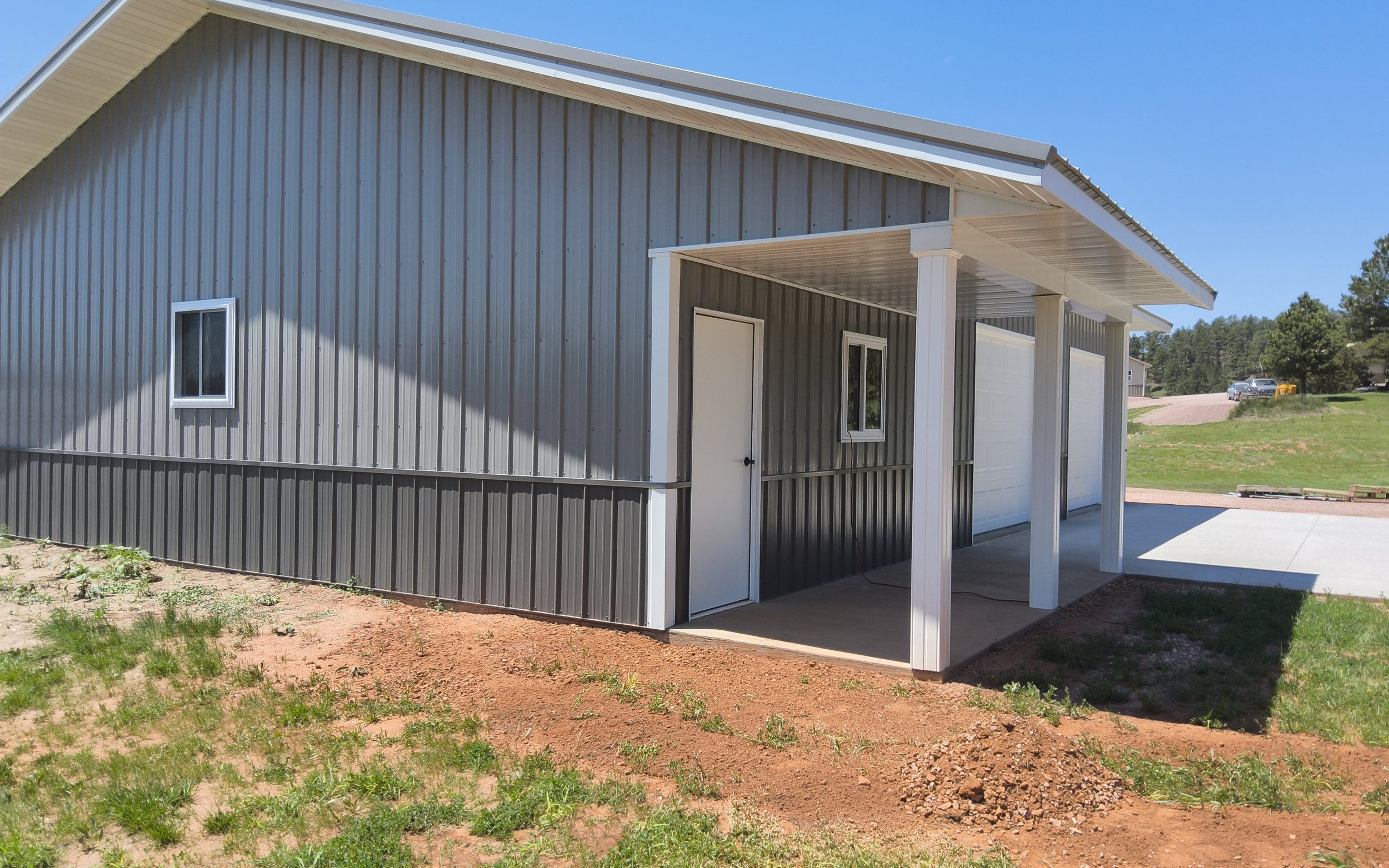 New gray metal garage with white trim and a covered porch area, set on a dirt and grass yard under a clear blue sky.