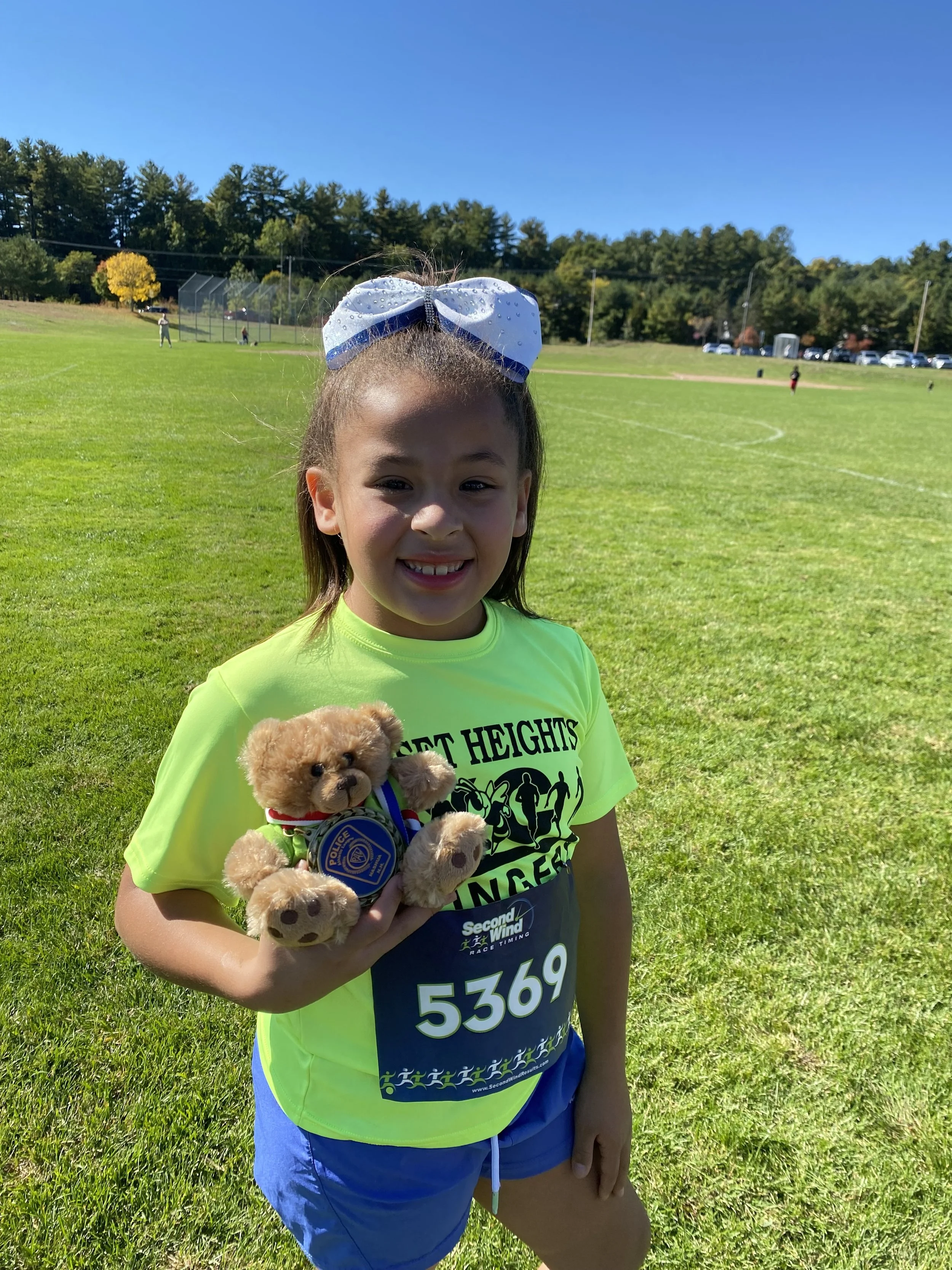 A girl in a neon yellow T-shirt with the words 'Great Heights Ultimate' holding a small teddy bear wearing a police badge, standing on a grassy field during a race or outdoor event.