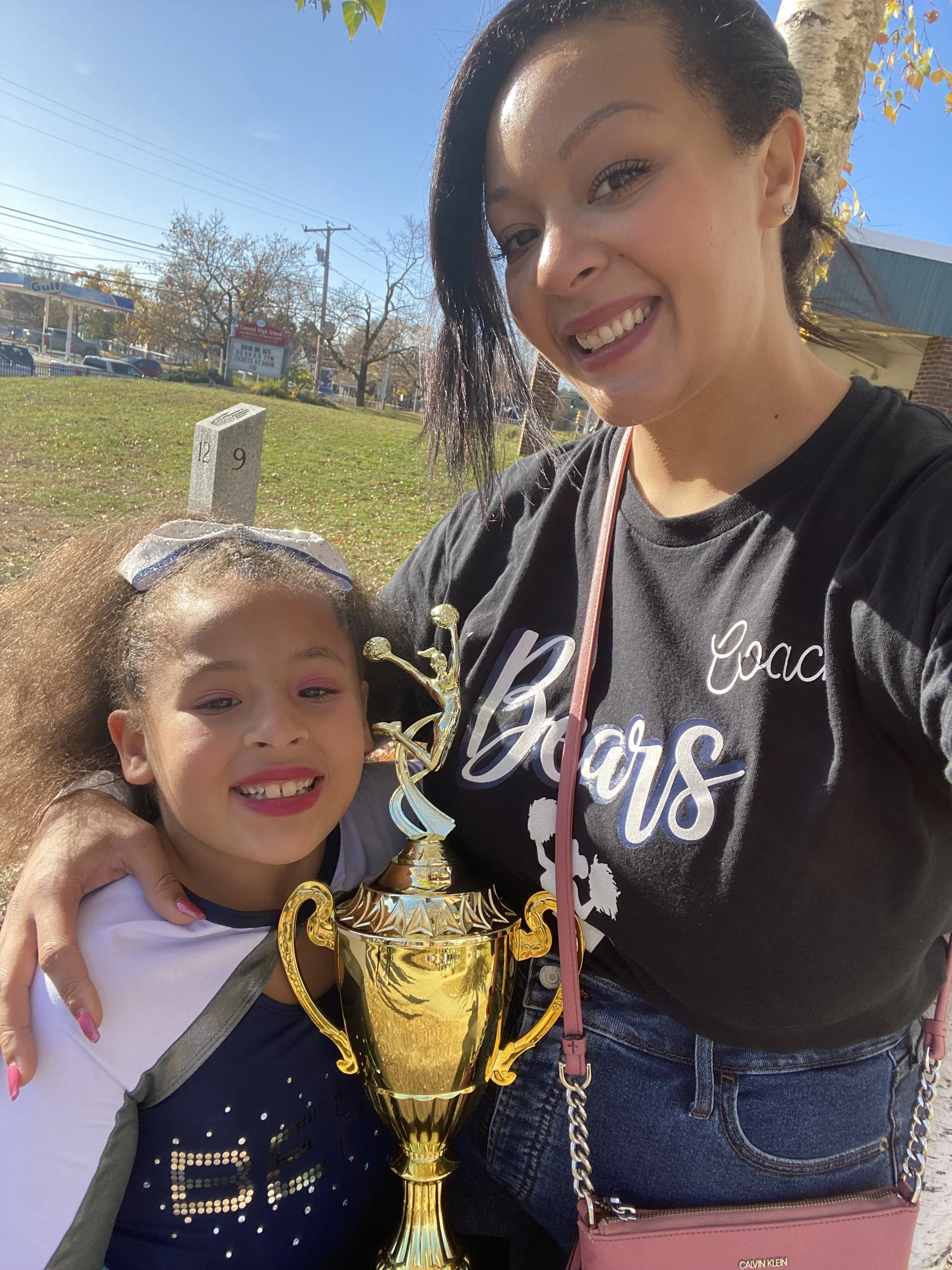 A woman and a young girl smiling outdoors, holding a gold trophy. The woman is wearing a black t-shirt with white writing, and the girl has a bow in her hair. The background has trees, a grass field, and a gas station.