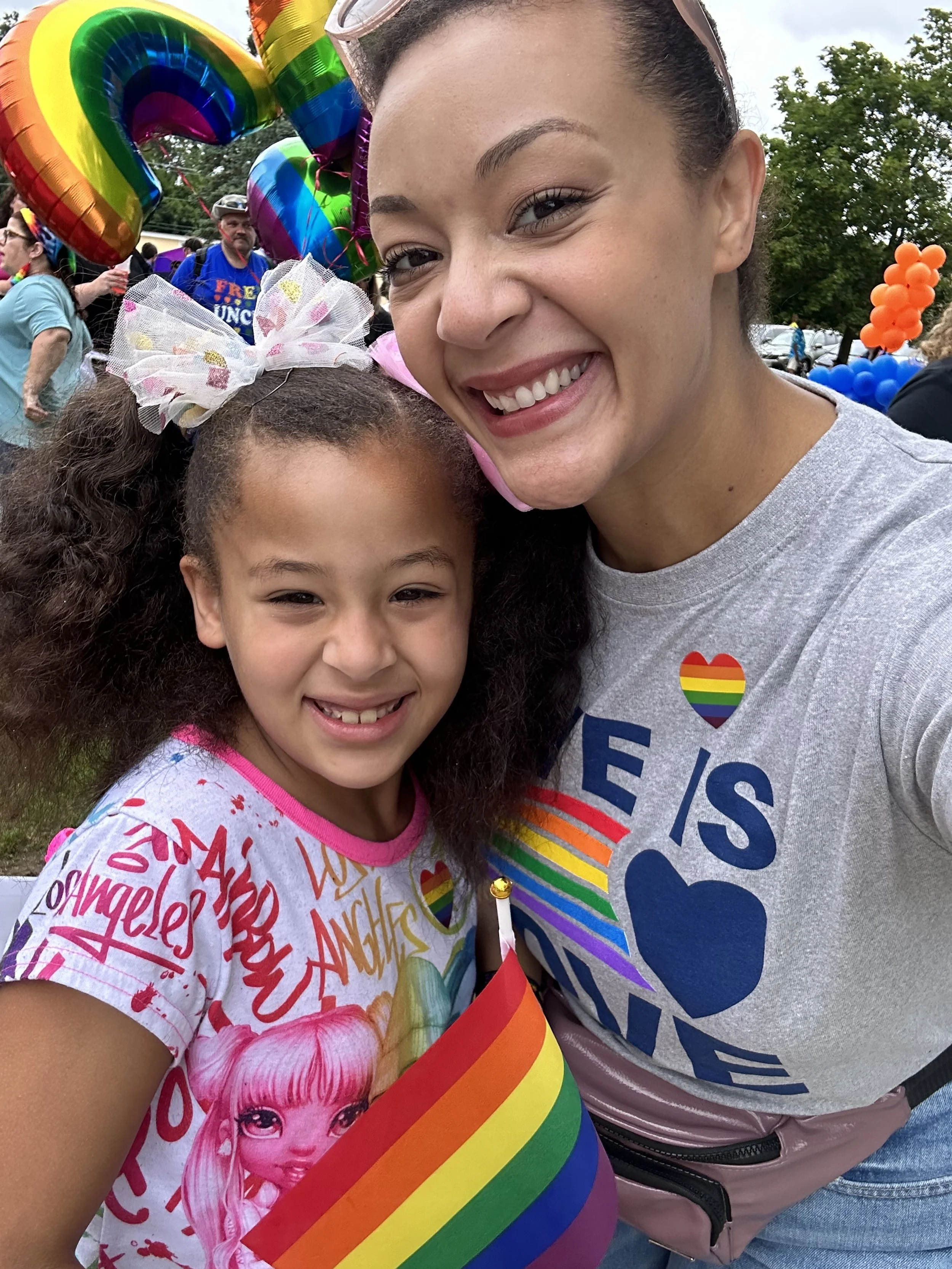 A smiling woman and girl at a pride parade, surrounded by rainbow balloons and flags, both wearing pride-themed clothing and accessories.