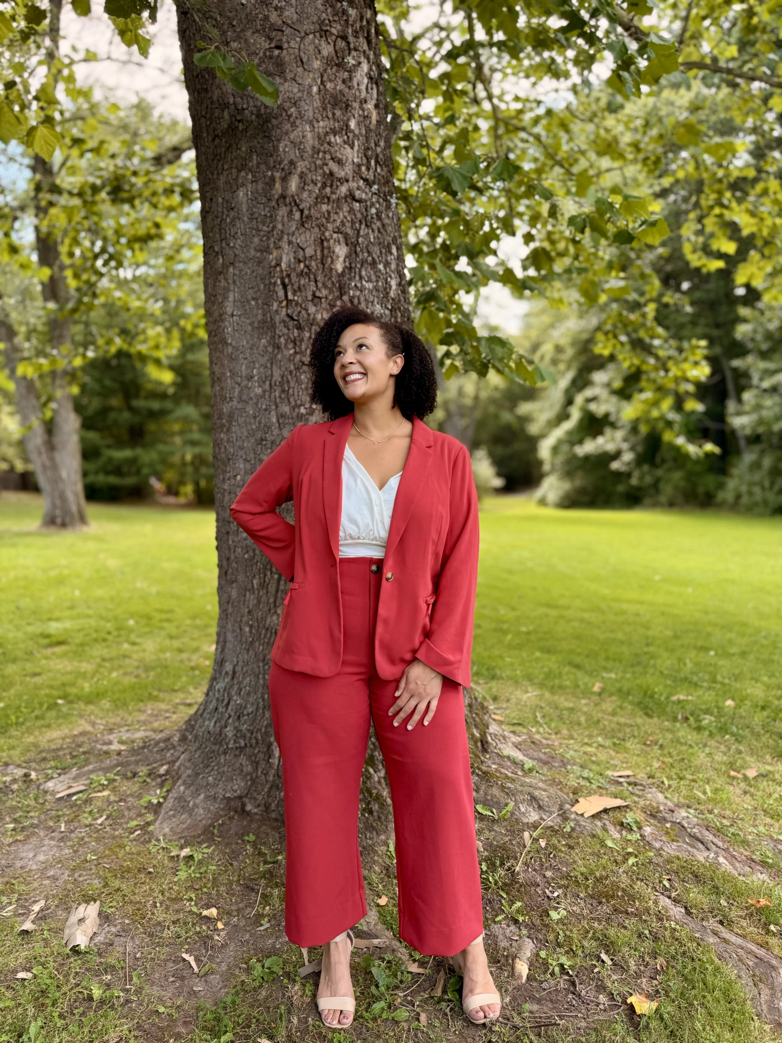 A woman in a red suit with beige open-toe shoes standing outdoors by a large tree with green leaves, smiling and looking upward, in a park setting.