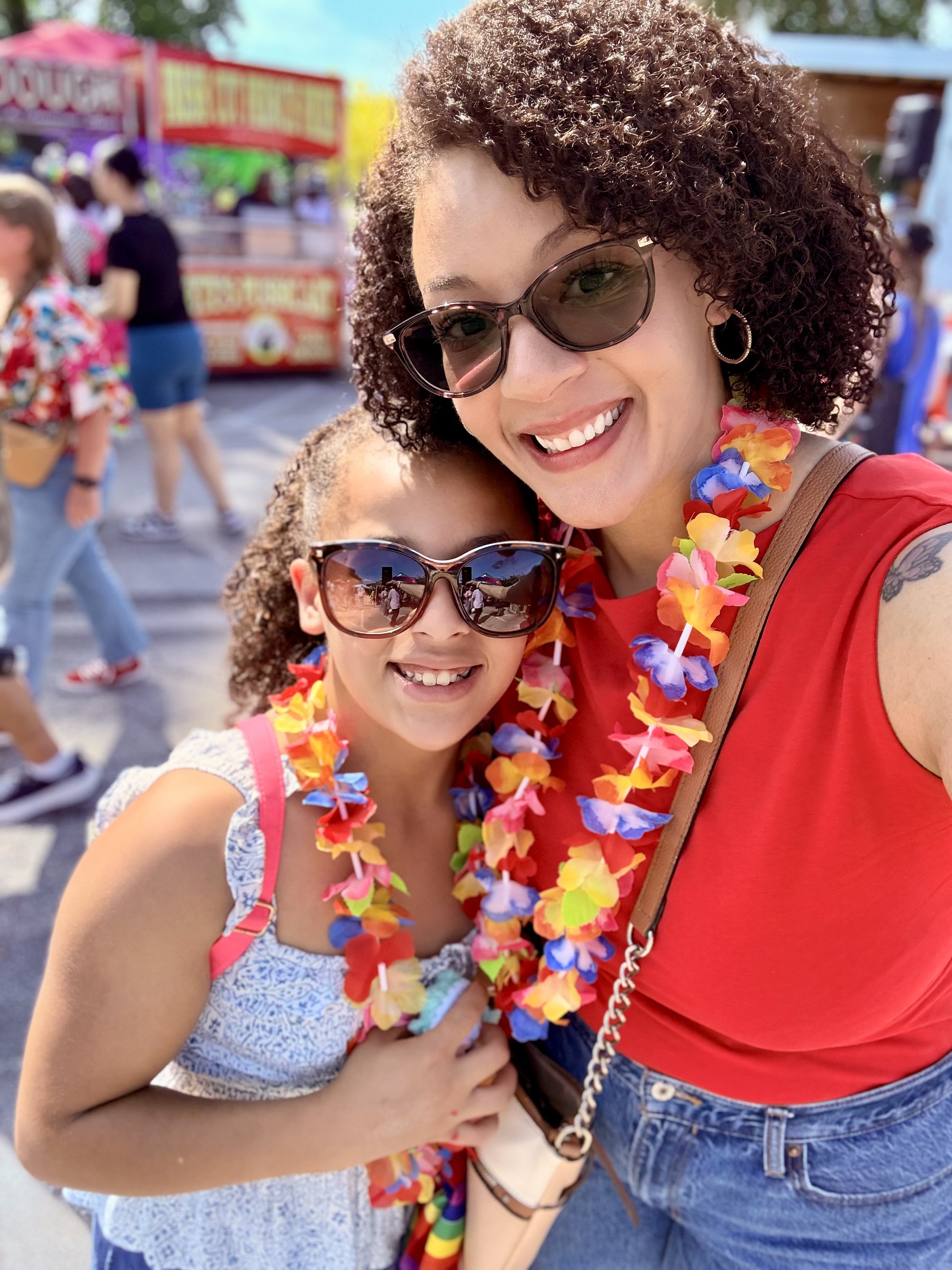 A woman and a young girl wearing sunglasses and colorful flower leis, smiling at the camera at an outdoor festival.