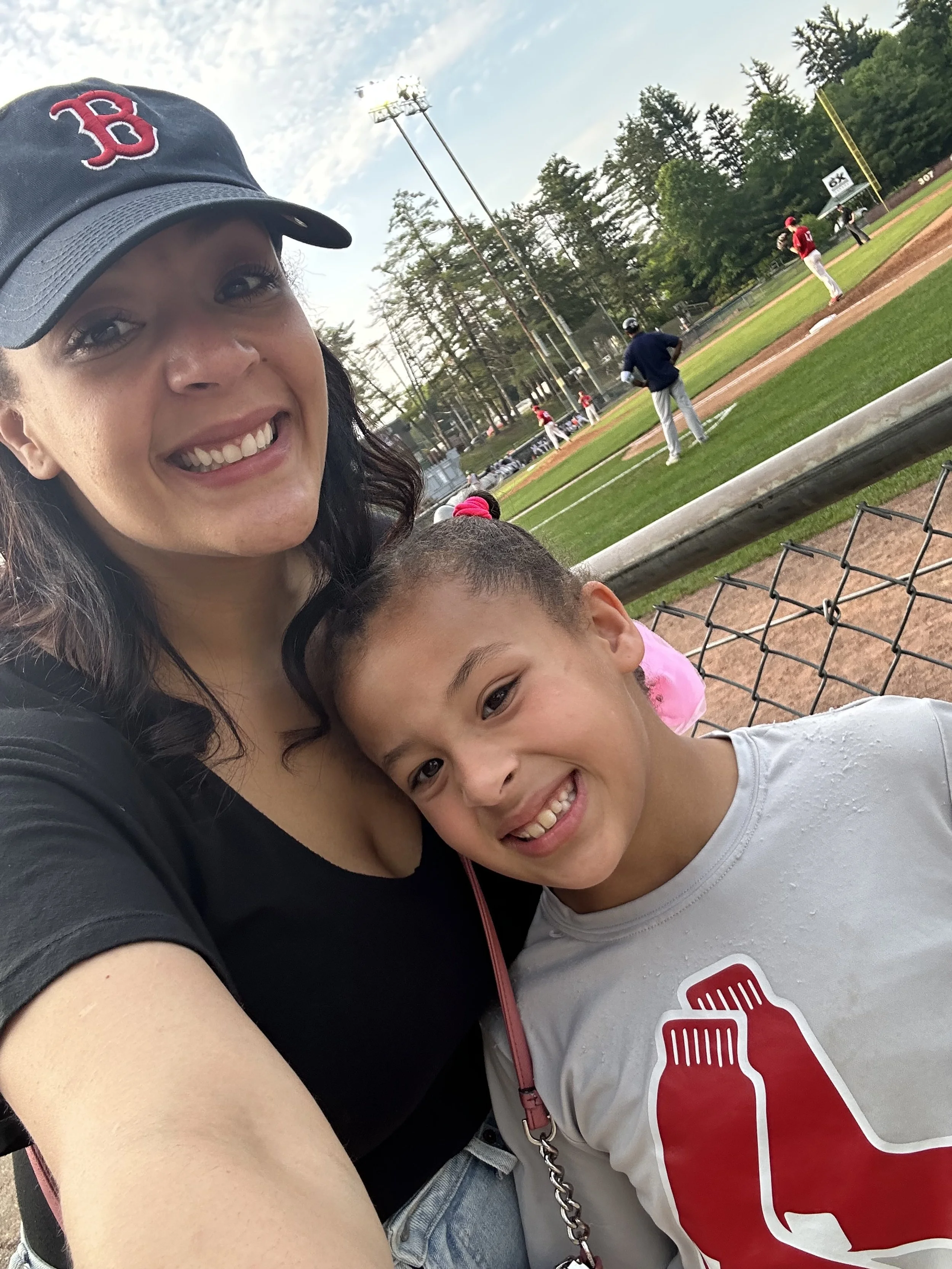 A woman and a girl taking a selfie at a baseball game, with players on the field in the background.