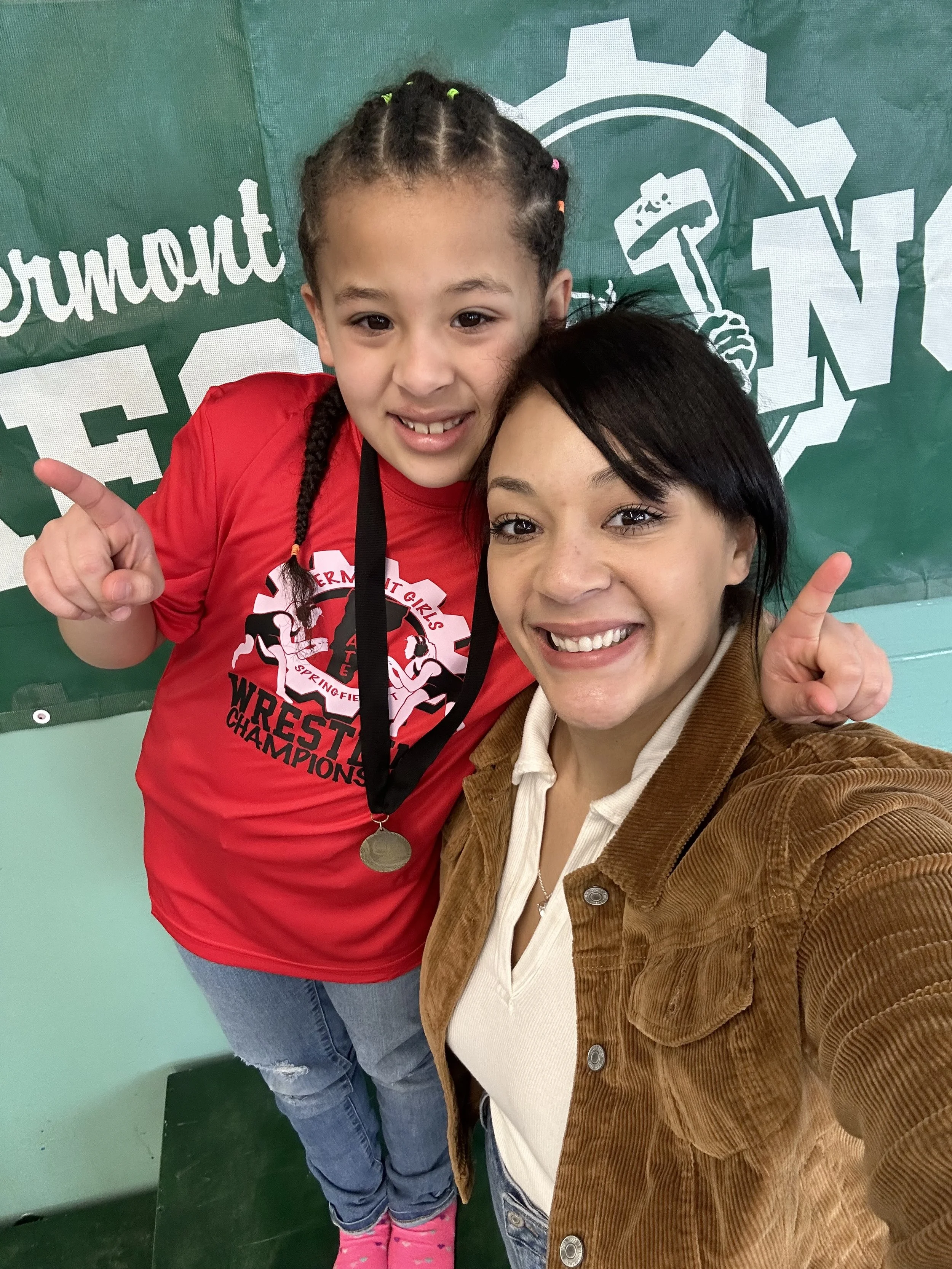 A young girl and a woman are smiling. The girl is wearing a red t-shirt with a wrestling graphic and a medal around her neck. They are standing in front of a green background with a gear and hammer graphic and the word 'Springfield' partially visible