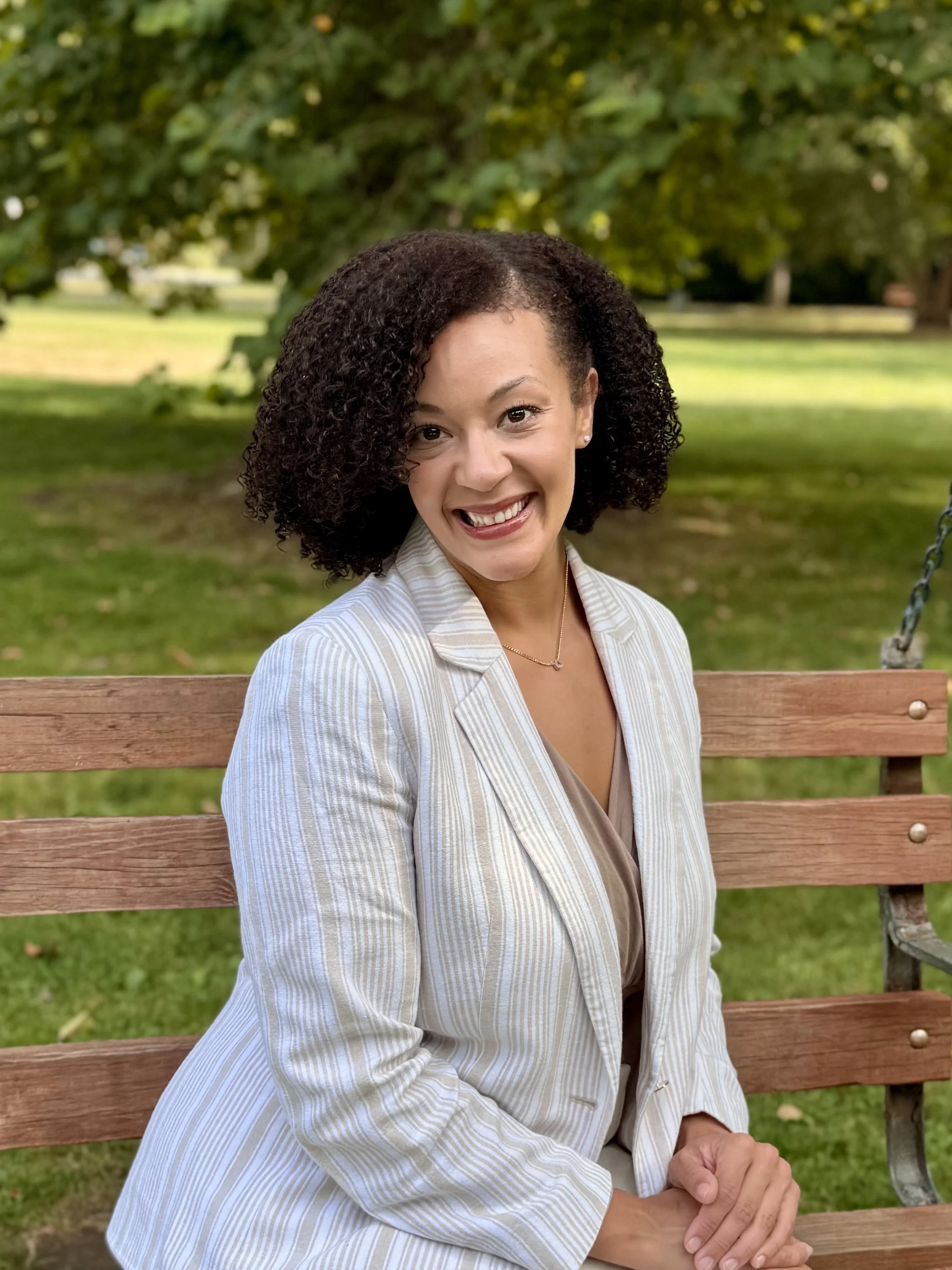 A woman with curly dark hair smiling while sitting on a wooden park bench with green trees and grass in the background.