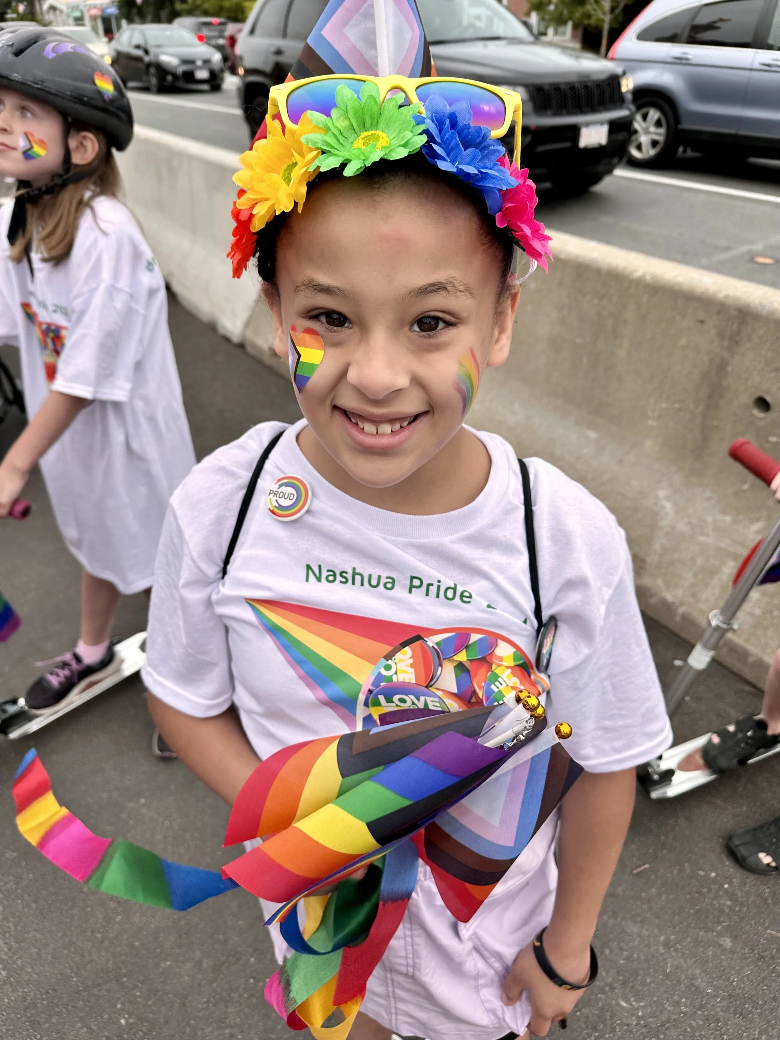 A young girl smiling at a pride parade, wearing rainbow stickers on her face and a white shirt with rainbow designs, holding rainbow-colored flags and wearing colorful flower headband and sunglasses.