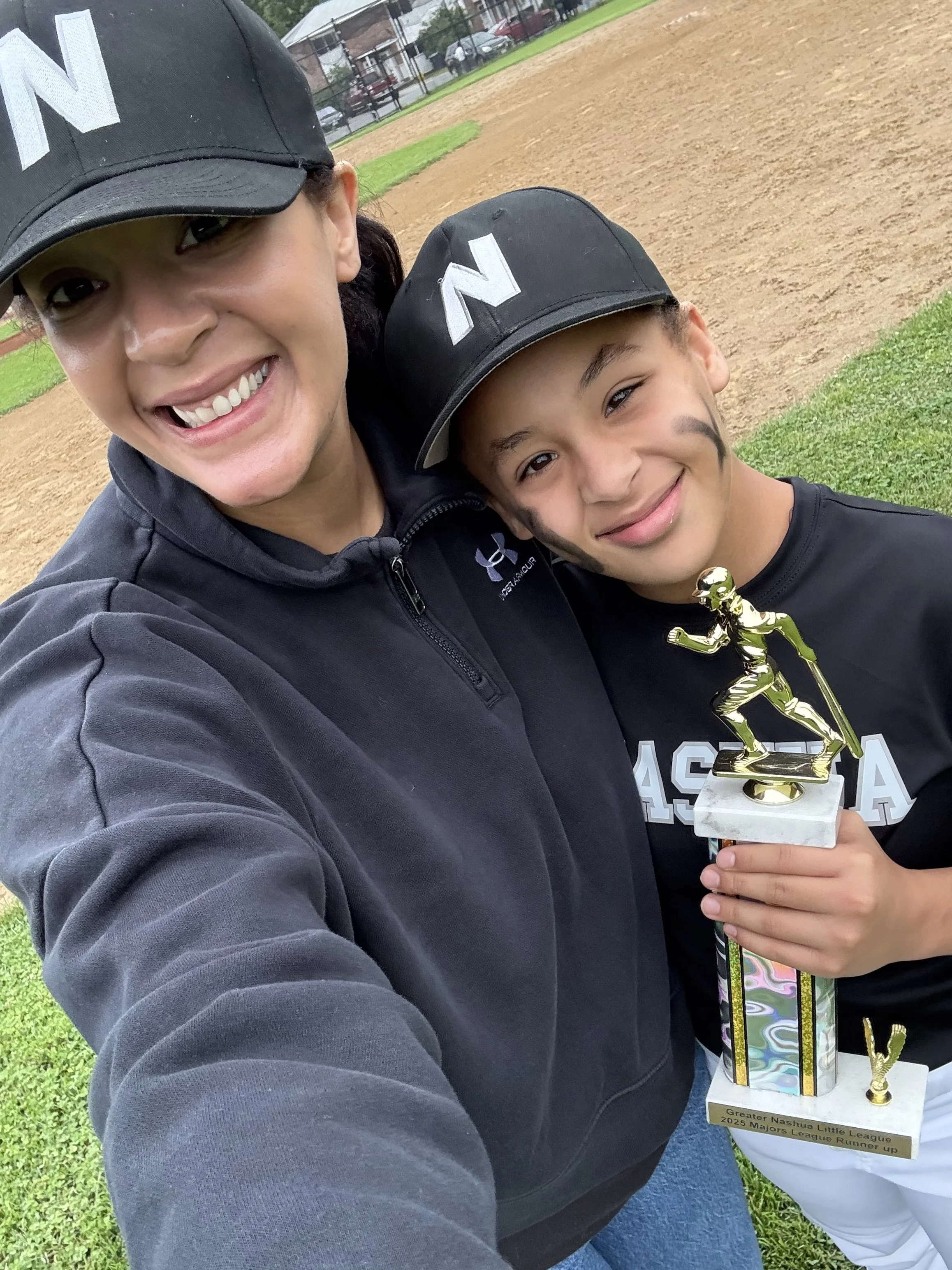 A woman and a young git; taking a selfie on a baseball field, with the girl holding a trophy. Both are wearing black caps and sports clothing, smiling at the camera.