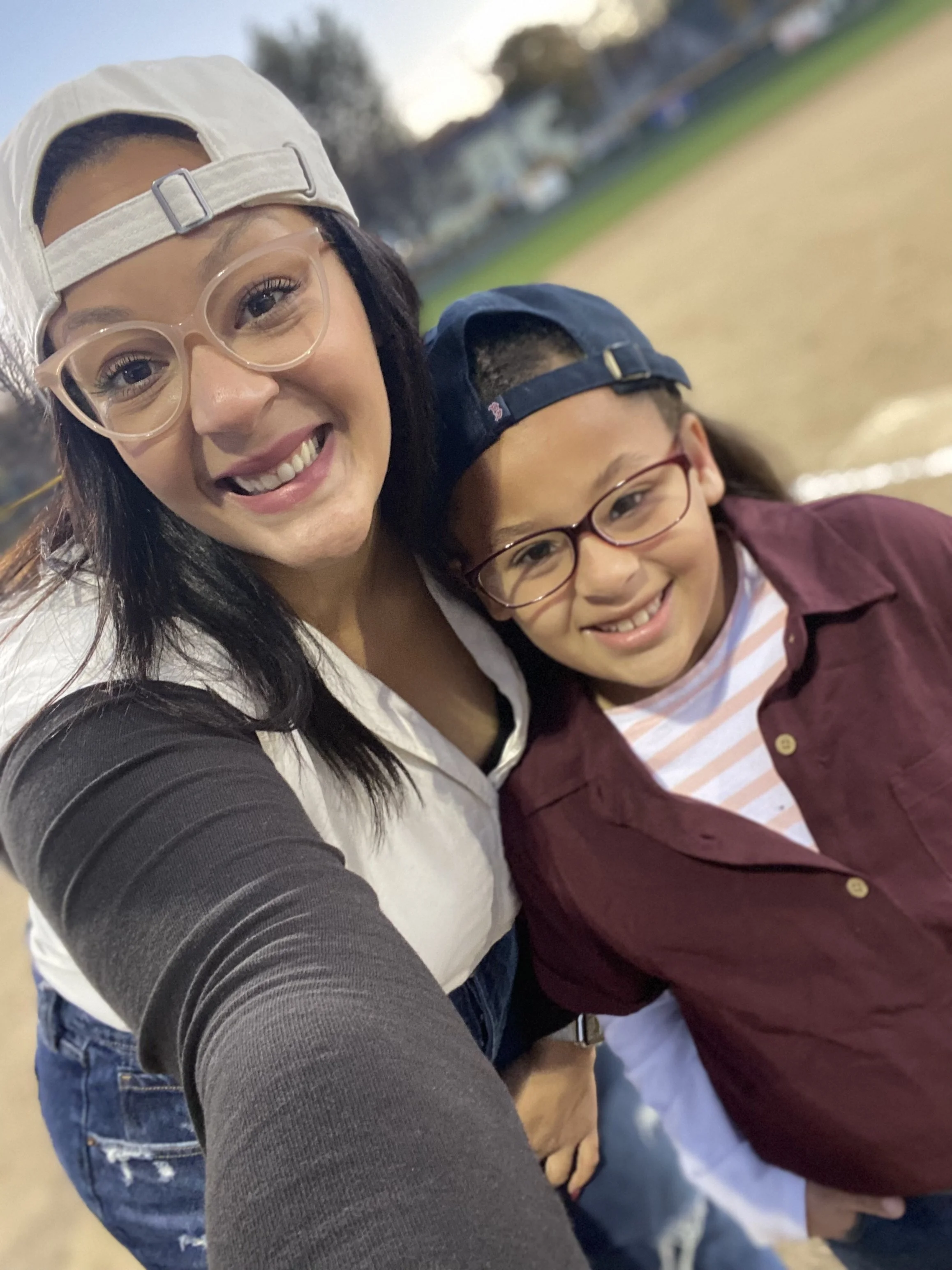 A woman and a girl taking a selfie together on a field during the daytime, both wearing caps and glasses, smiling at the camera.