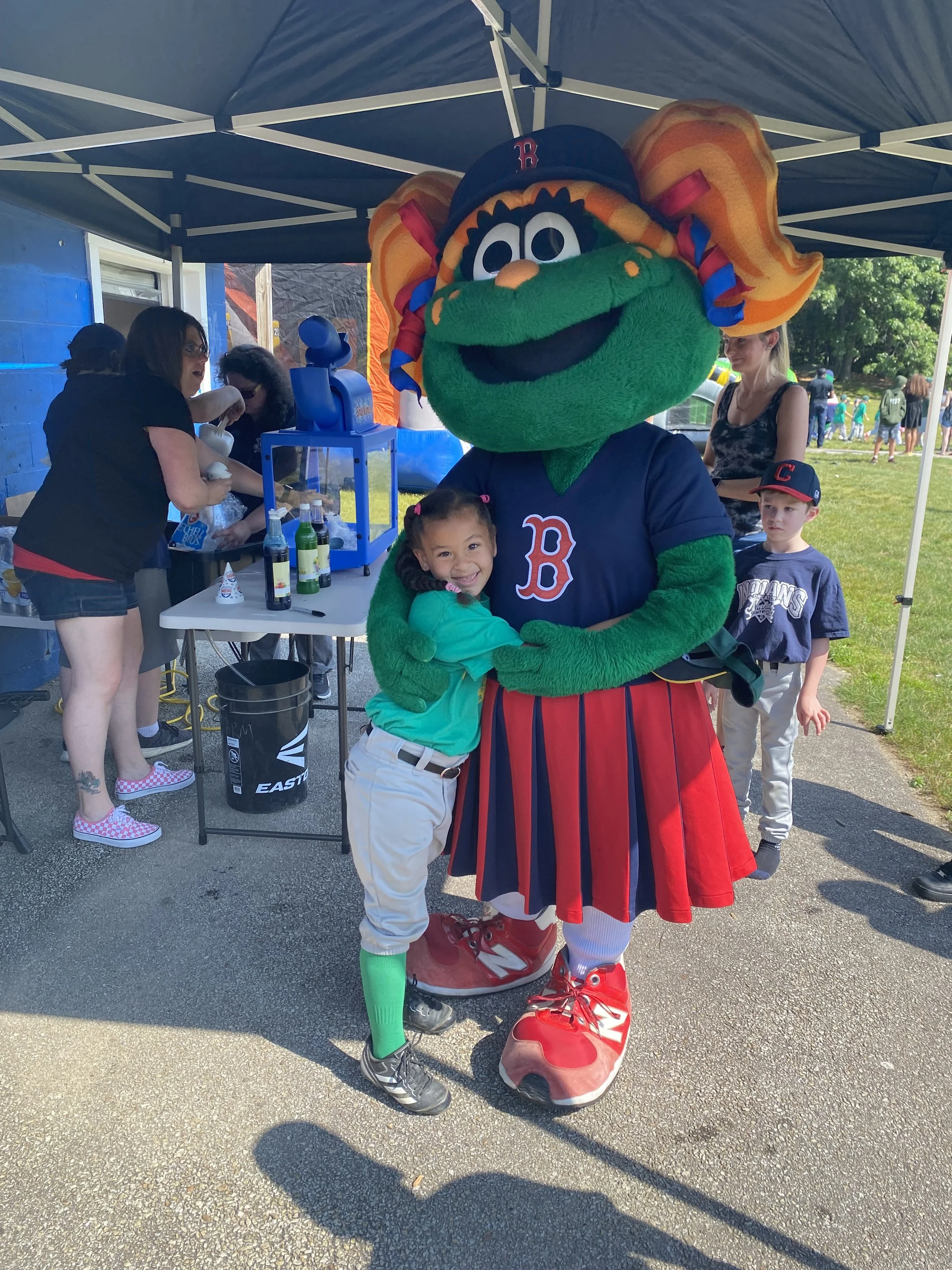 A young girl hugging a Red Sox mascot dressed as a colorful, green character with orange hair, a navy jersey with a red 'B', a red and navy skirt, and large red shoes at an outdoor event under a canopy.