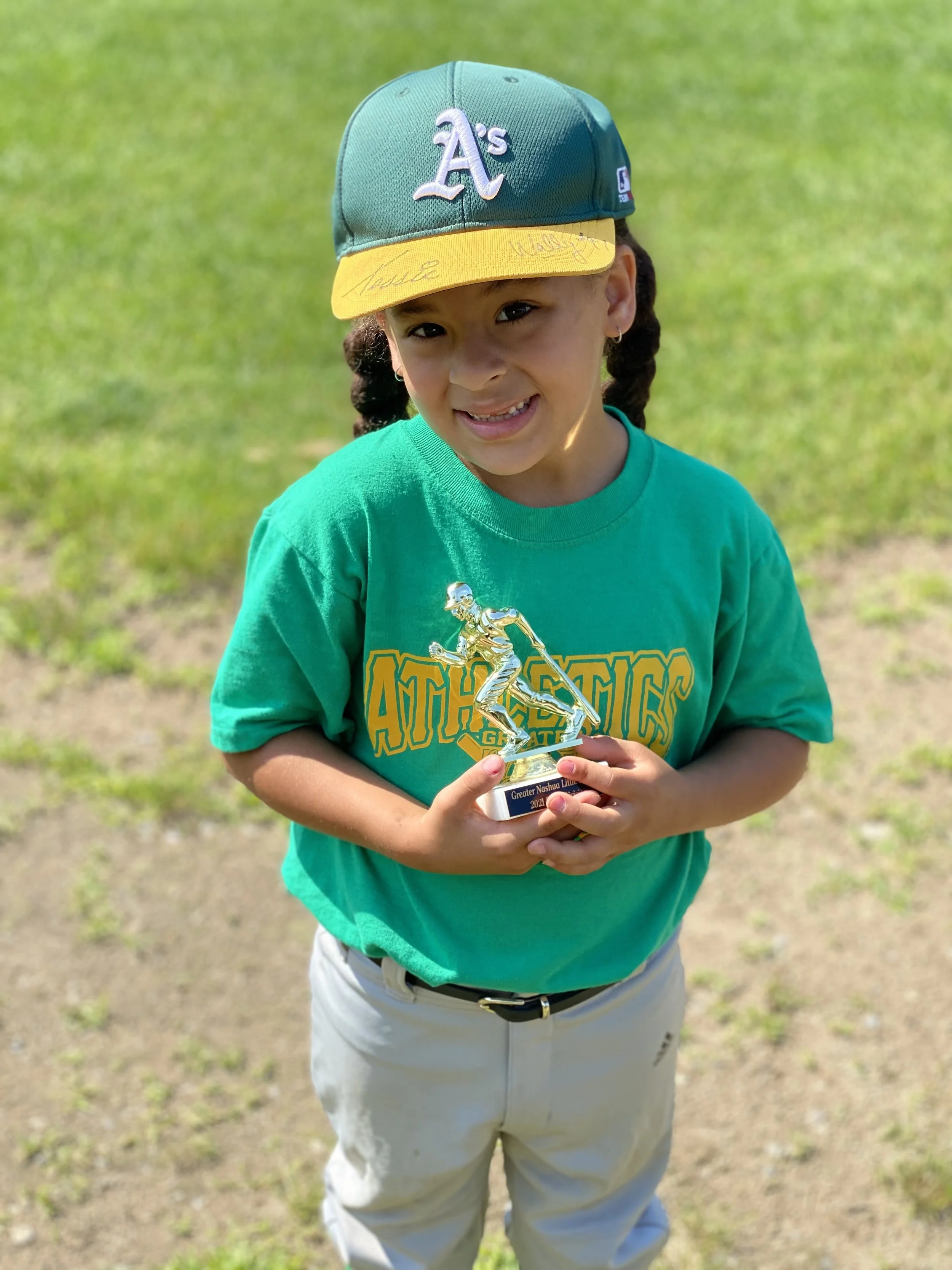 Young girl with braided hair wearing a teal t-shirt and a teal and yellow baseball cap, holding a small trophy, standing outdoors on grass.