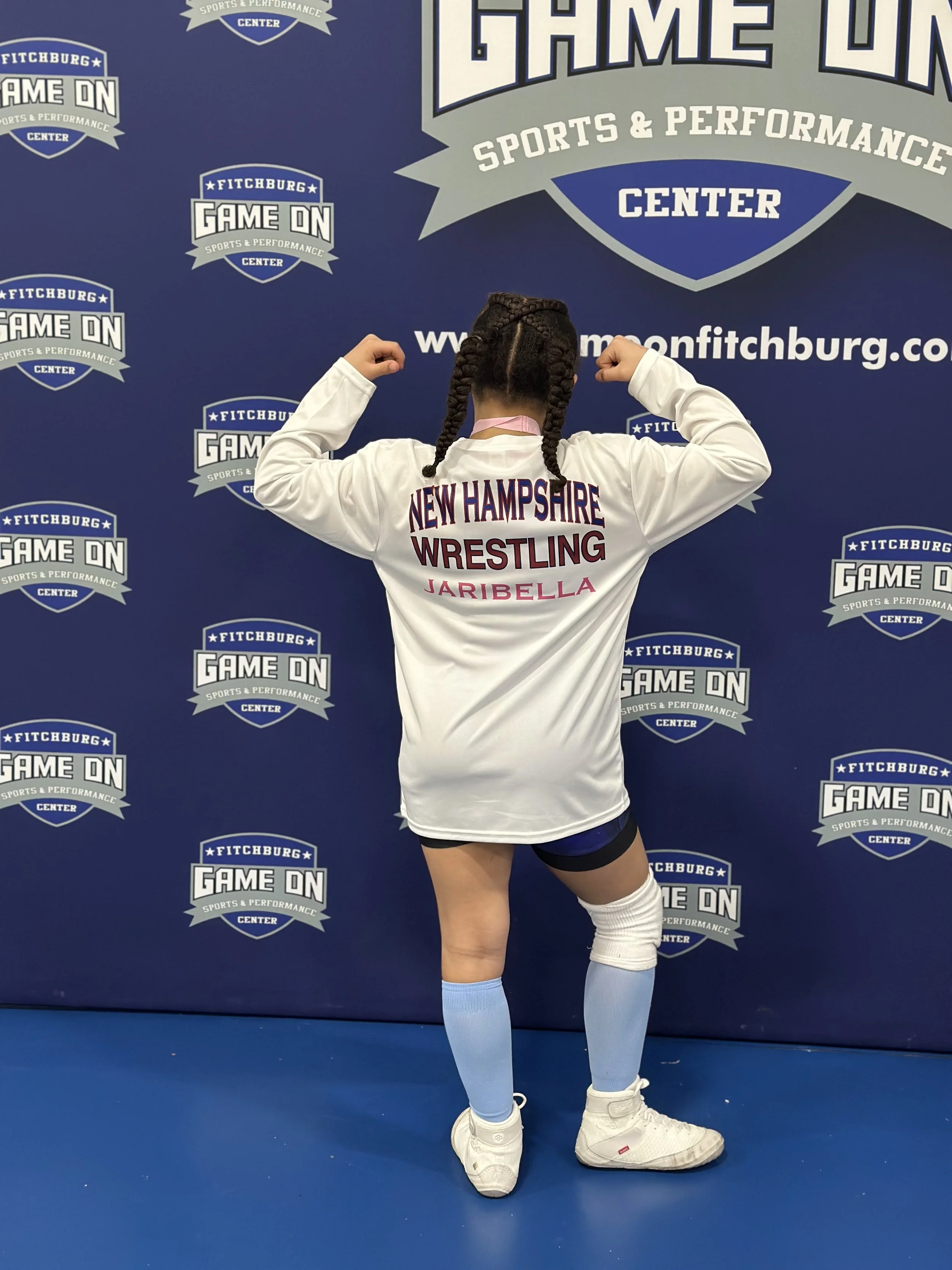A female athlete with braided hair poses with her back to the camera, flexing her arms, in front of a blue backdrop with logos and text for Fitchburg Game On Sports & Performance Center. She is wearing a white shirt with 'New Hampshire Wrestling'.