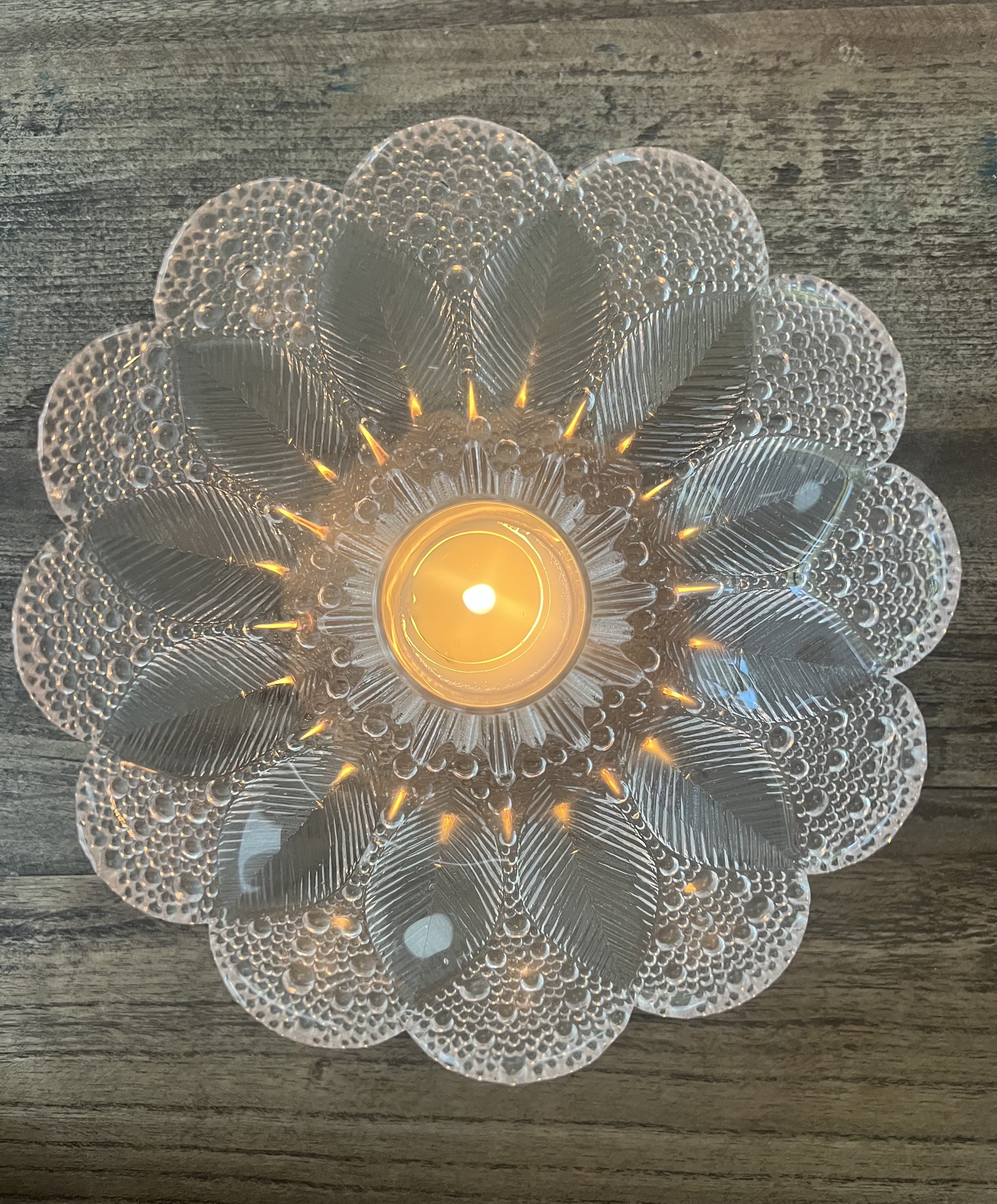 Decorative glass candle holder with a lit candle inside, designed to resemble a flower with petal-shaped glass pieces, on a wooden surface.