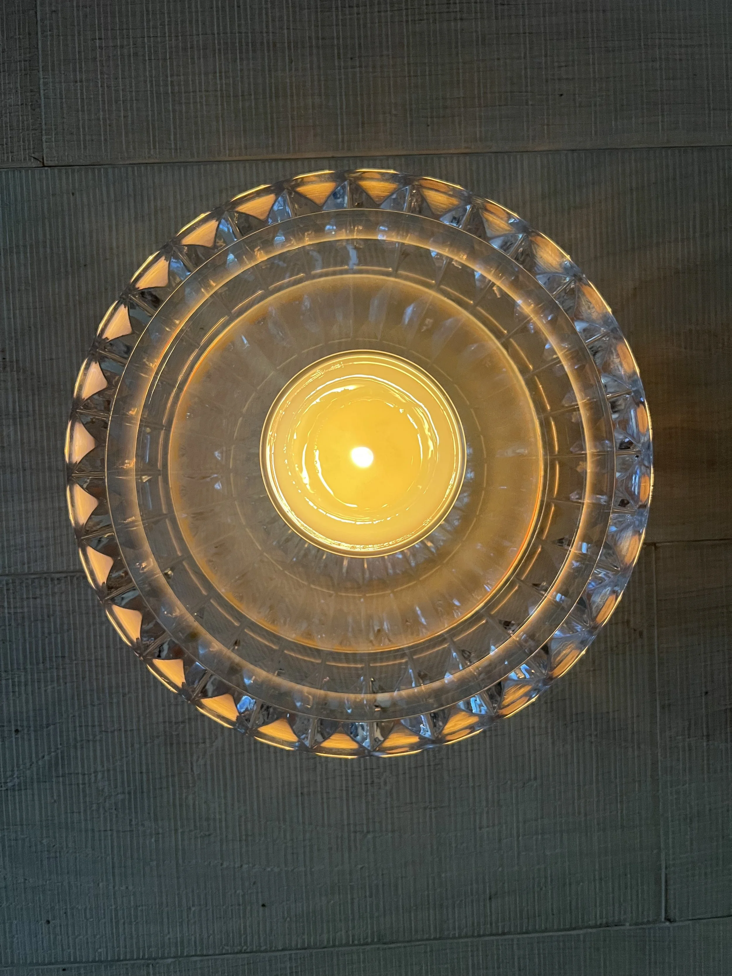 Overhead view of a ceiling light fixture with a glowing yellow bulb, surrounded by a decorative glass or crystal ring, attached to a wood-paneled ceiling.