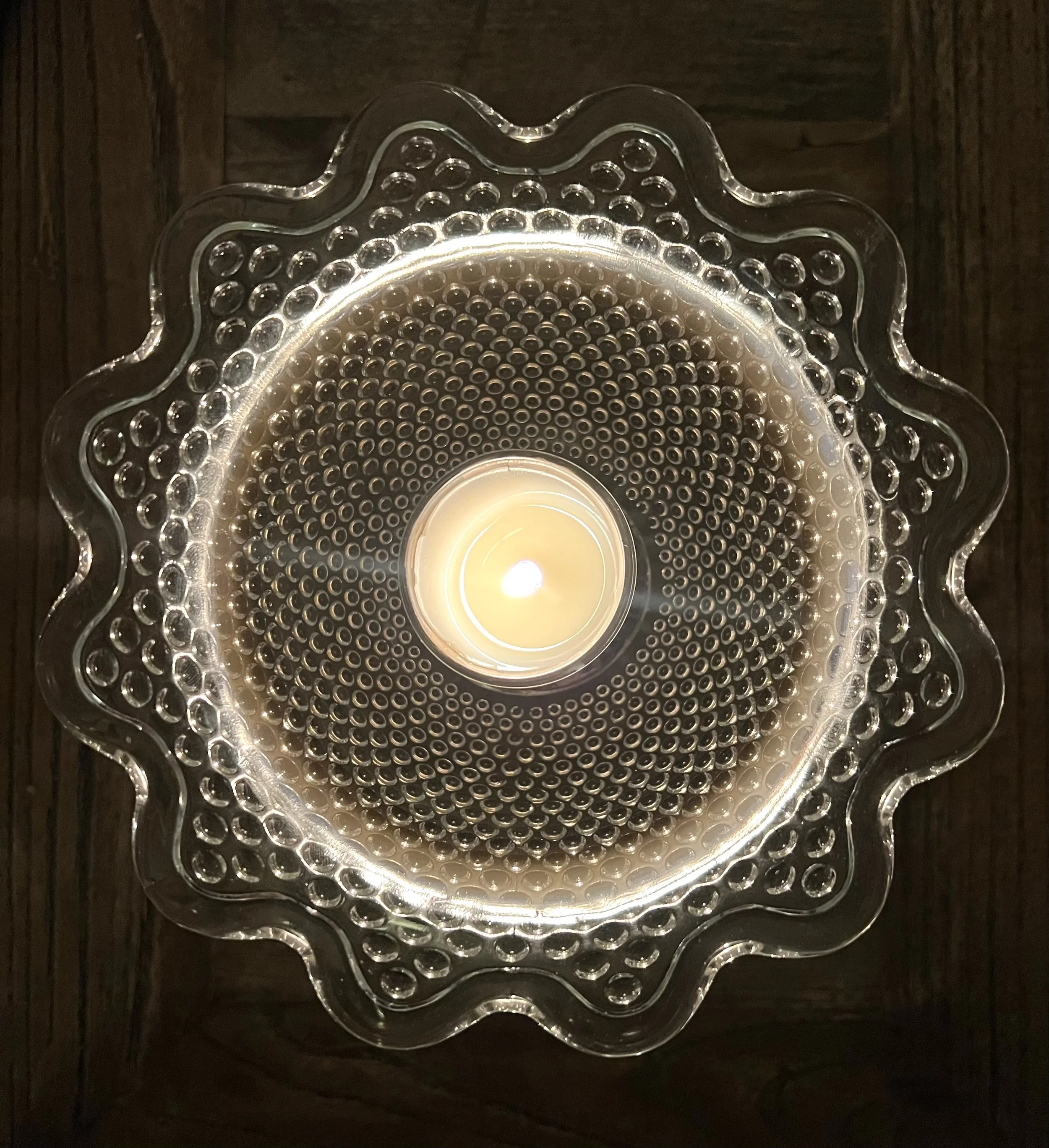 A decorative glass chandelier with a circular pattern and scalloped edges, illuminated with a warm light bulb at the center, mounted on a dark wooden ceiling.