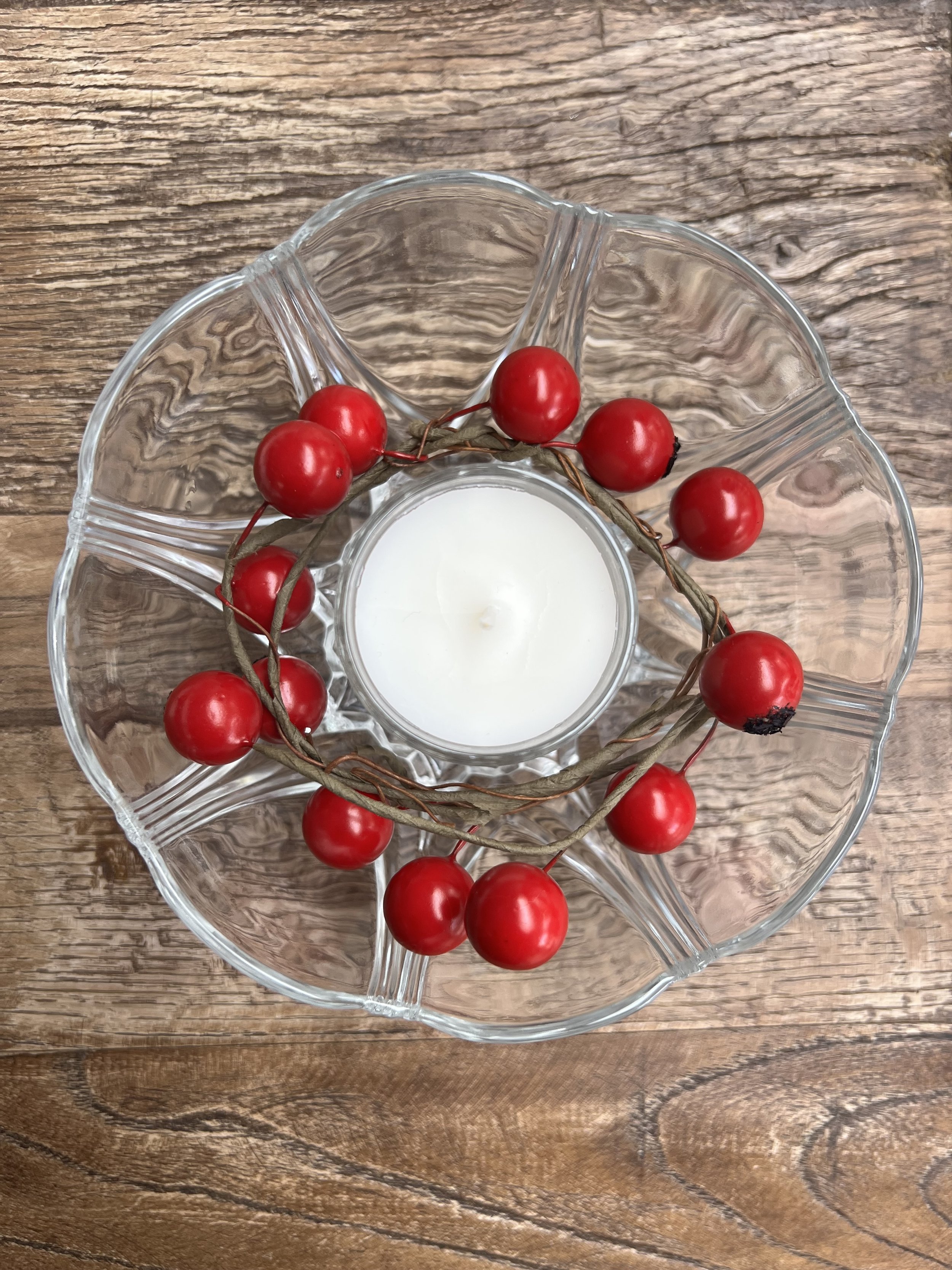 A glass candle holder with a white candle, surrounded by a wreath of red berries on a wooden surface.