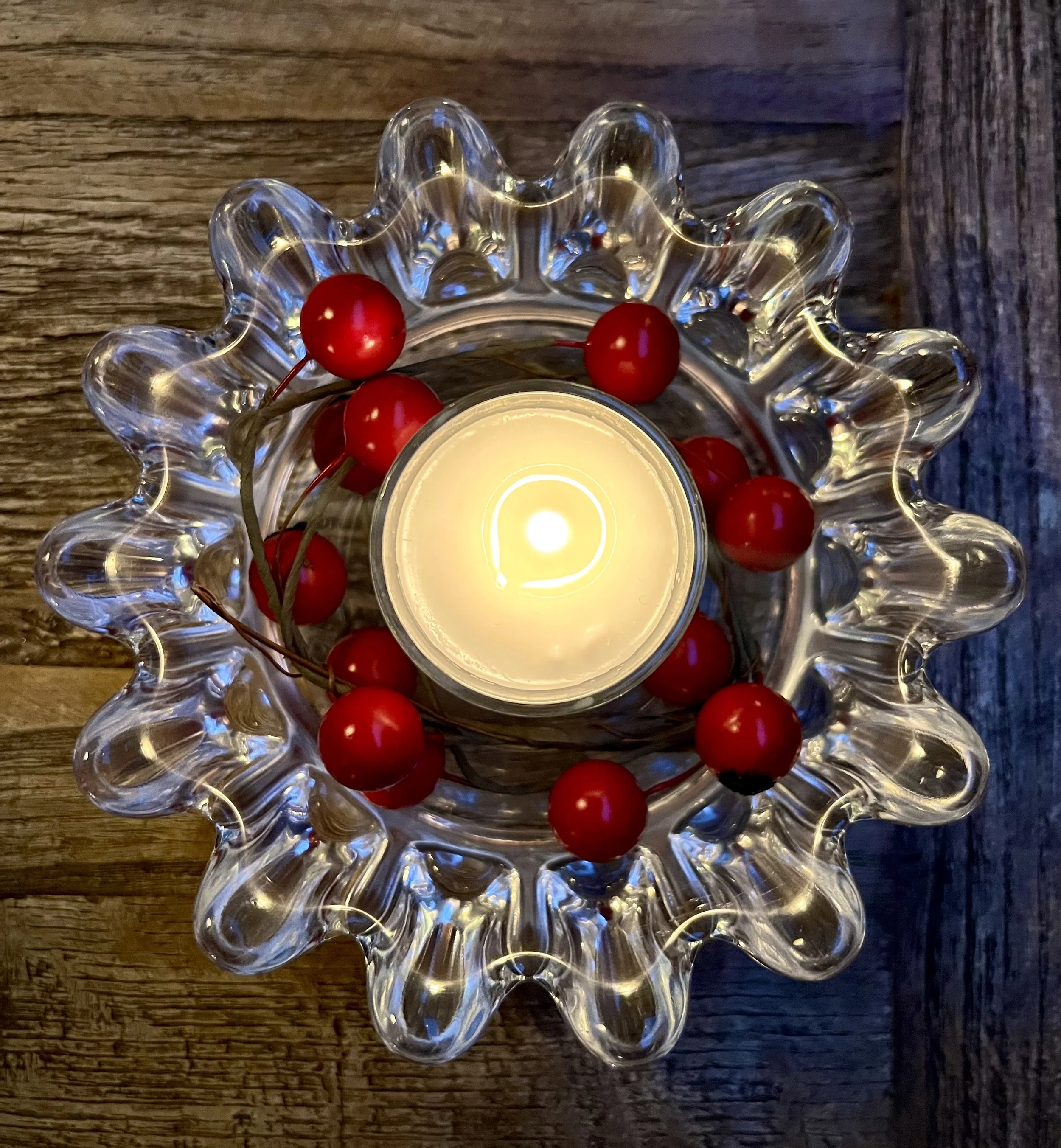A lit candle in a glass holder surrounded by a string of red berries in a glass dish on a wooden surface.