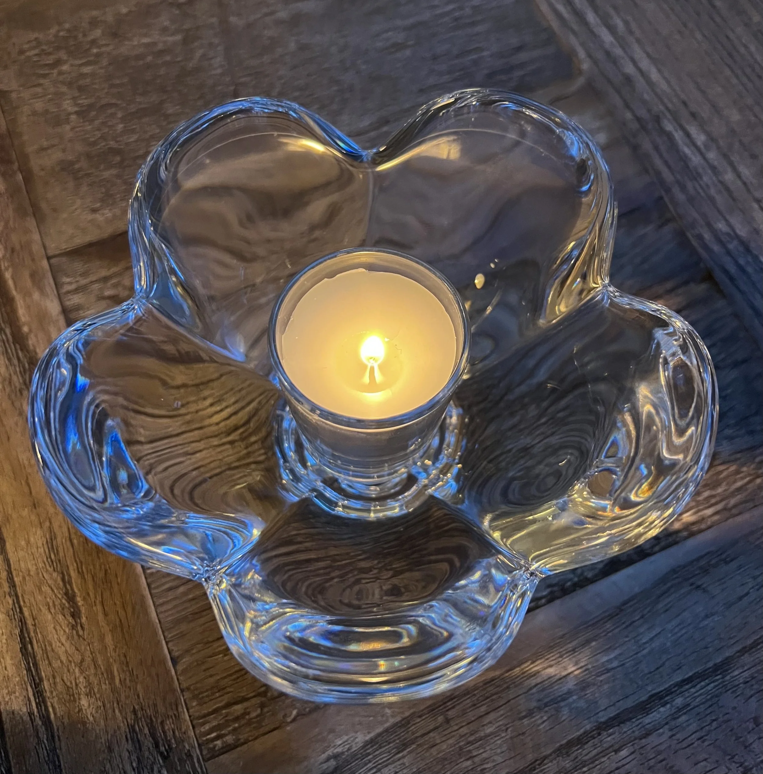 A lit candle in a glass holder placed inside a decorative glass bowl with wavy edges, on a wooden surface.