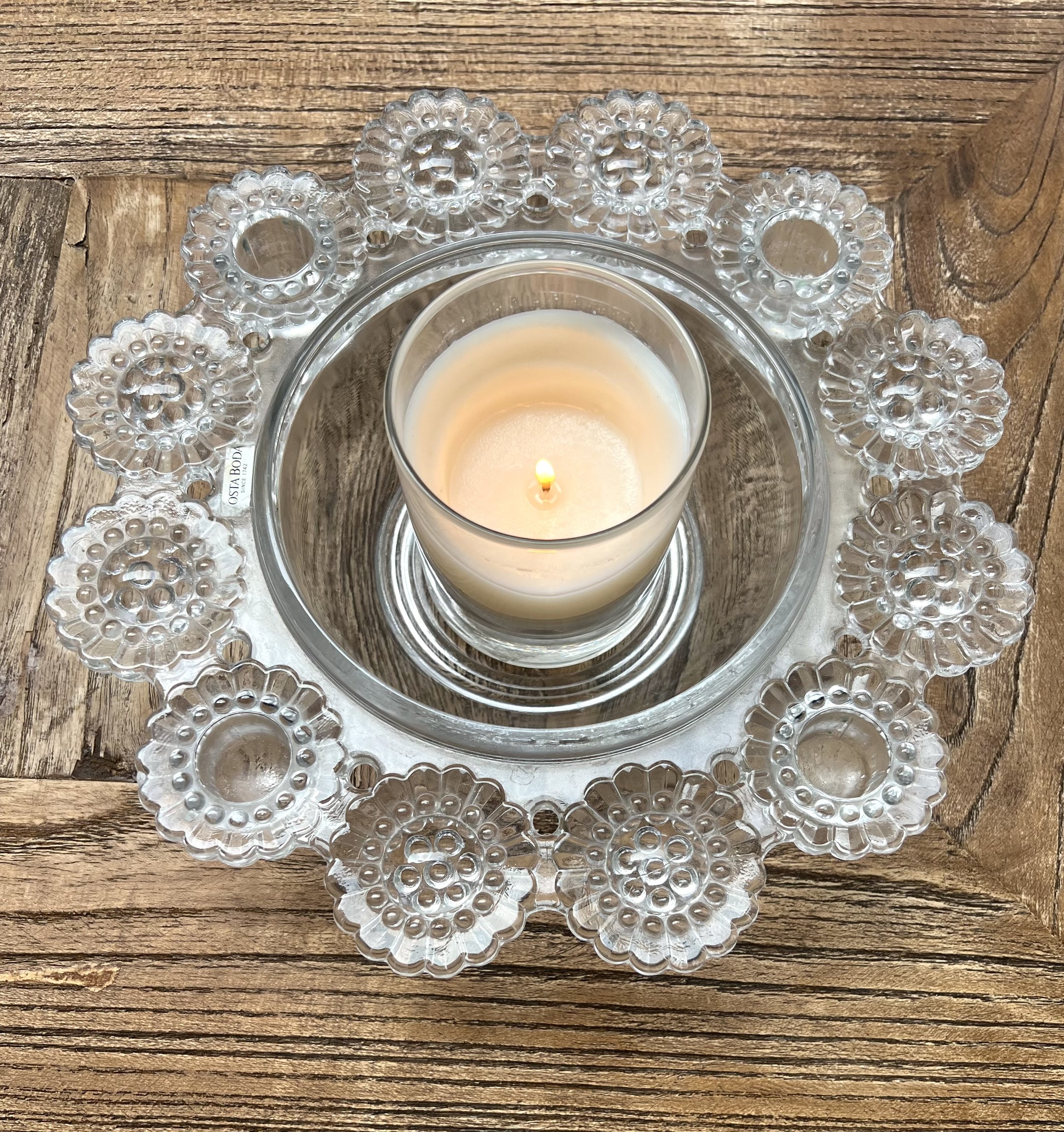 A lit white candle in a glass holder placed on a clear glass plate with decorative textured edges, on a wooden surface.