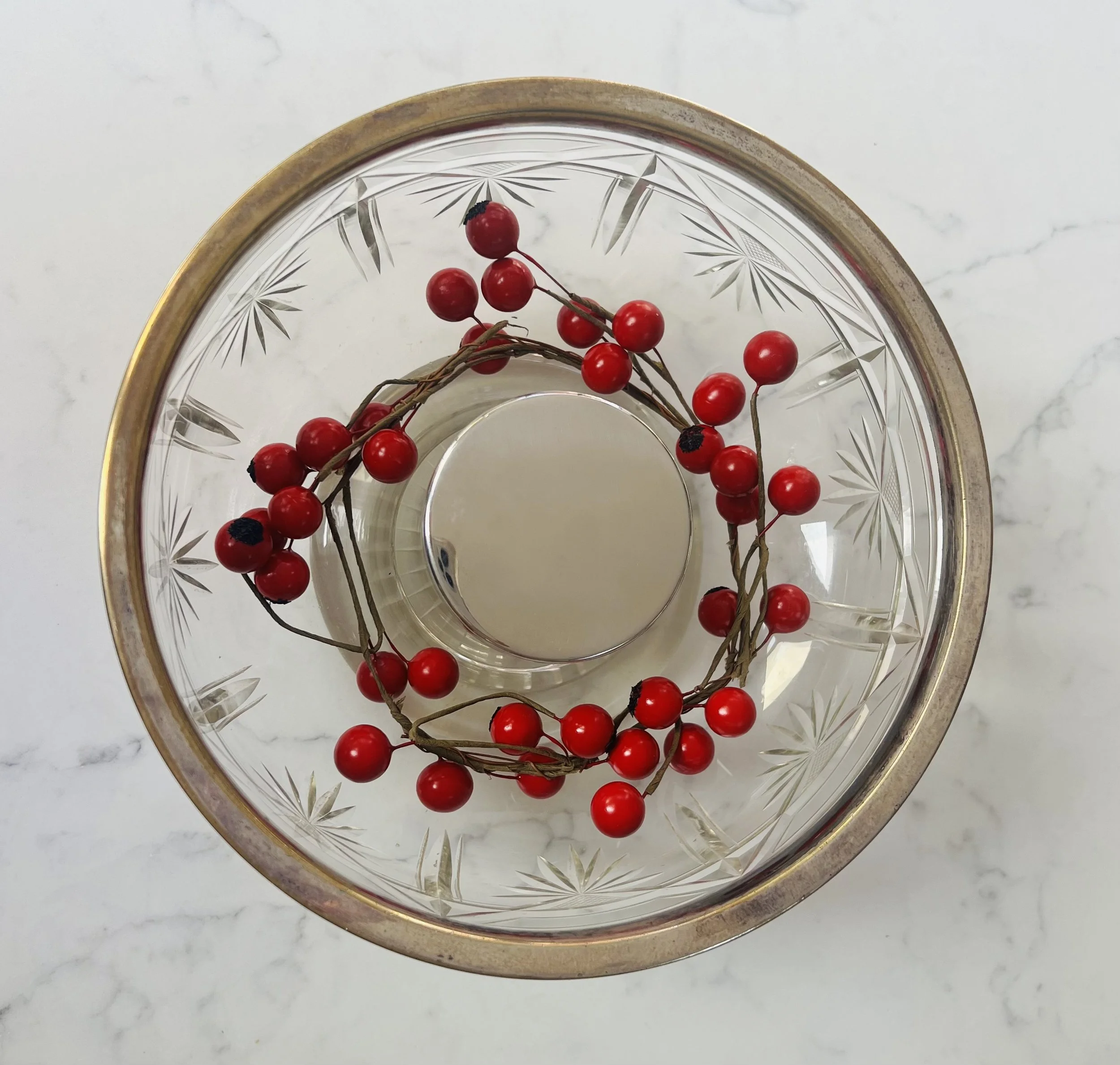 Top view of a glass bowl with etched starburst patterns, containing a string of red berries, on a white marble surface.