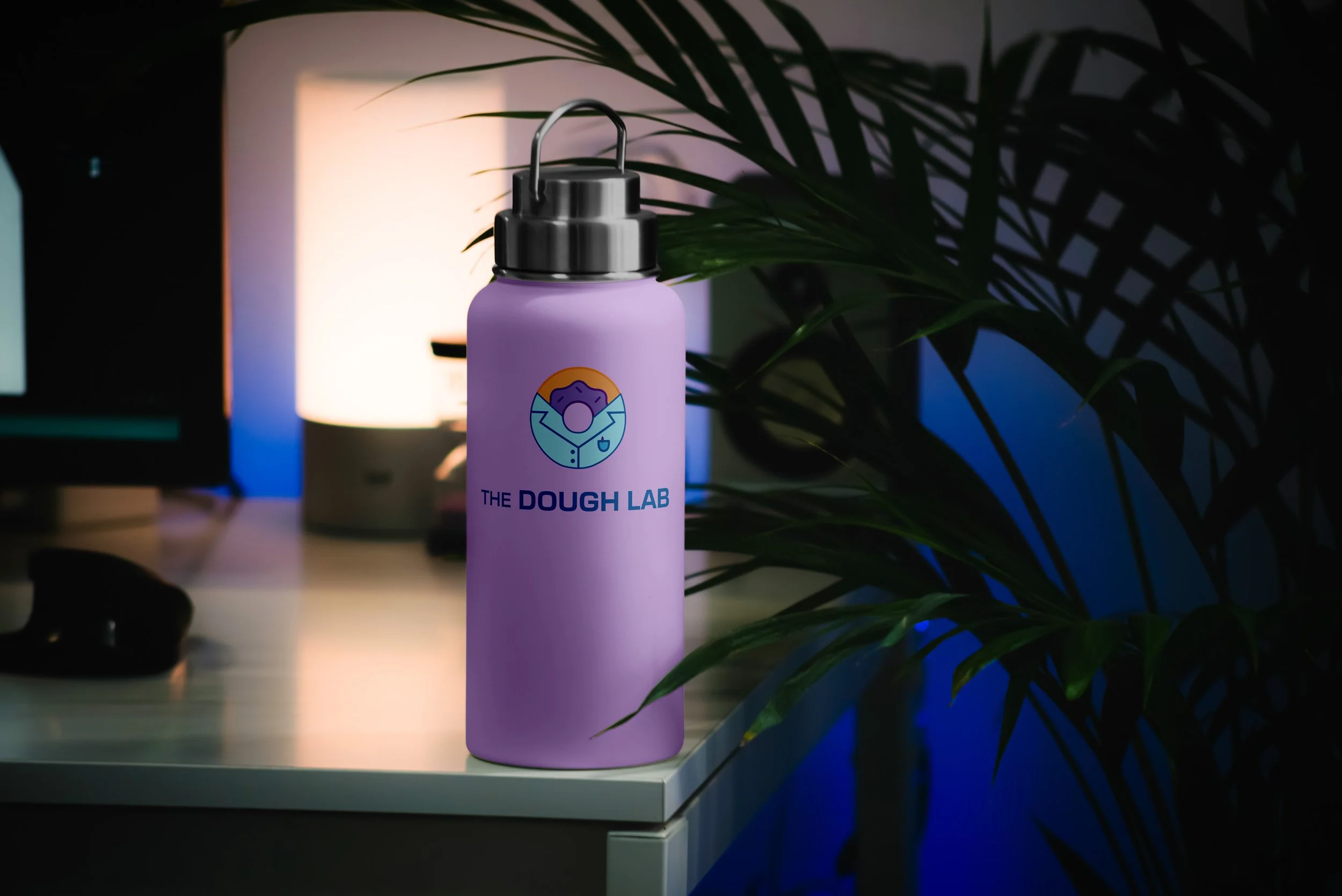 A purple water bottle with 'The Dough Lab' logo on a white table, next to a large green plant and illuminated by a computer monitor and ambient lighting.