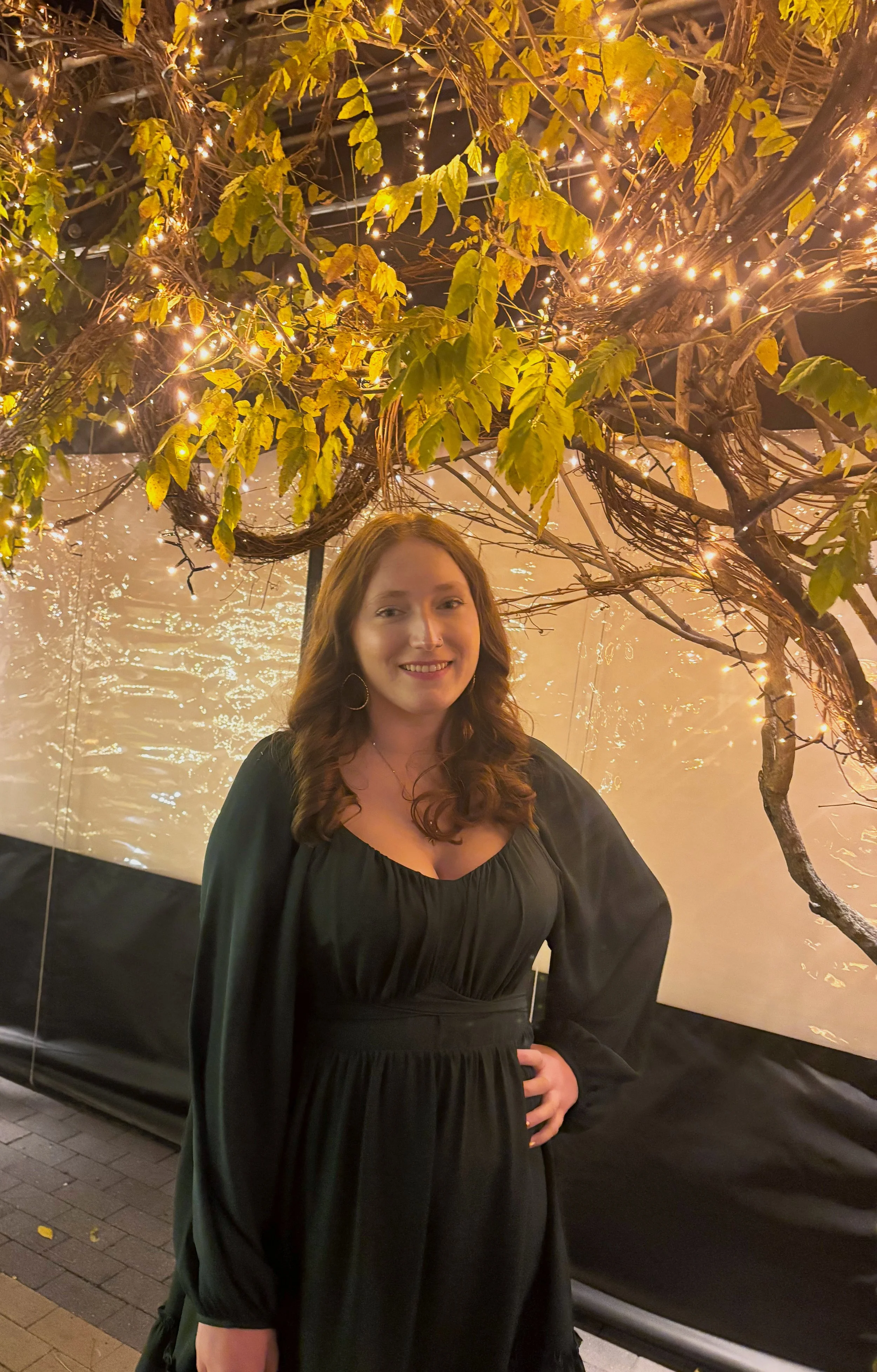 A woman with brown curly hair wearing a black dress standing in front of a backyard with string lights and a tree with yellow-green leaves.