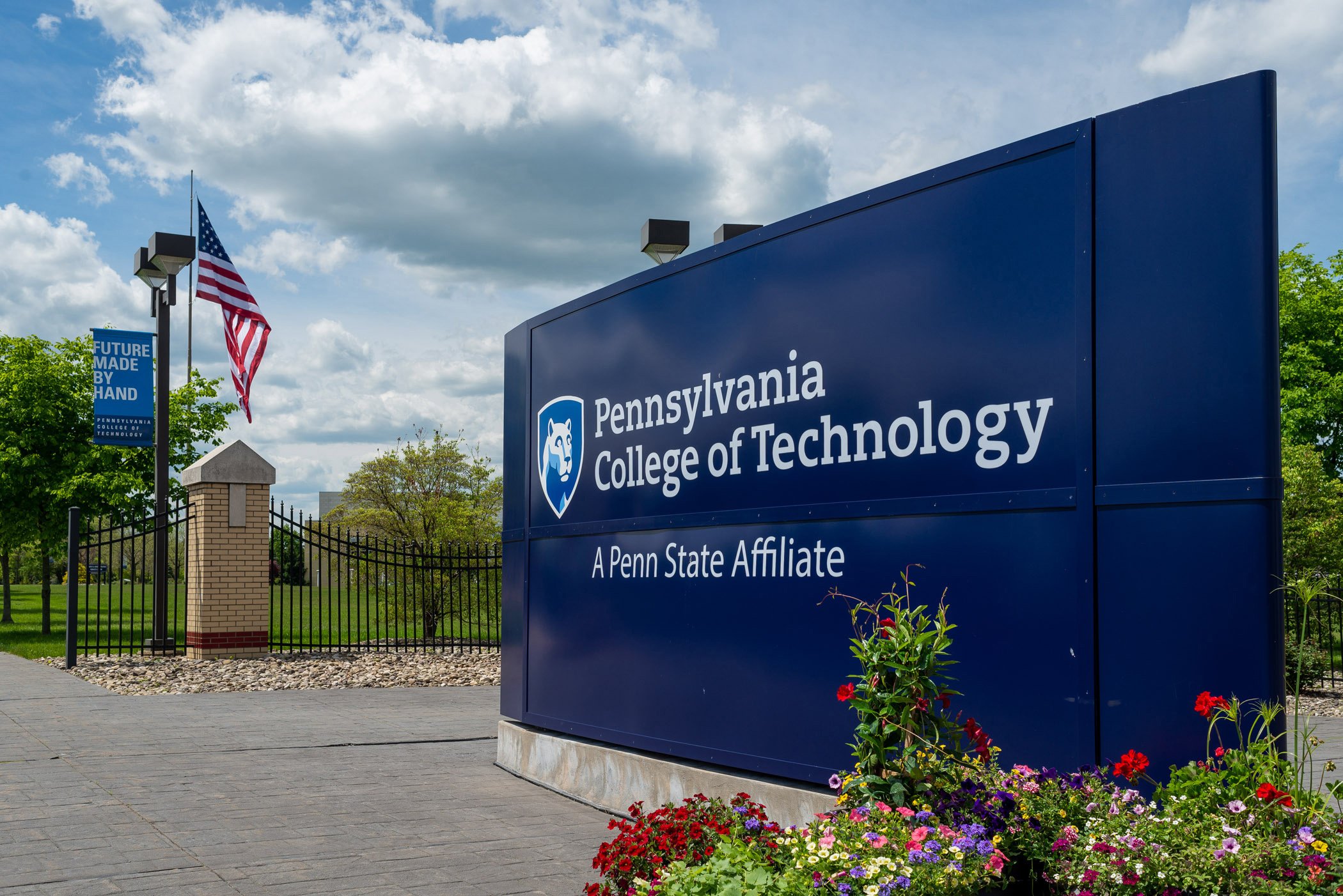 University sign for Pennsylvania College of Technology with flowers in the foreground, American flag, and a banner that reads "Future Made by Hand," outside on a cloudy day.