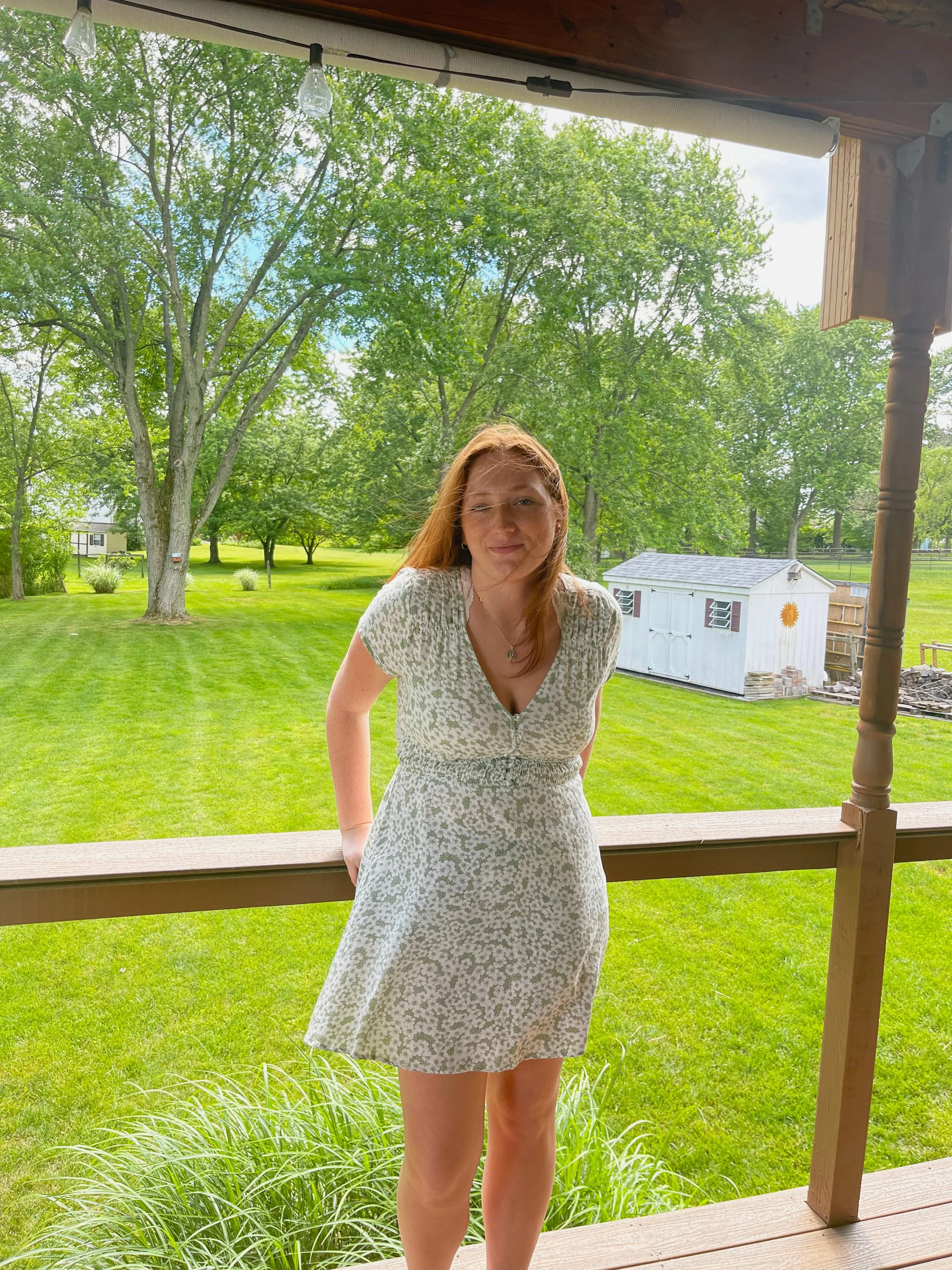 A woman with red hair outdoors on a porch, wearing a light-colored dress with a floral pattern, with a green backyard, trees, and a small white shed in the background.