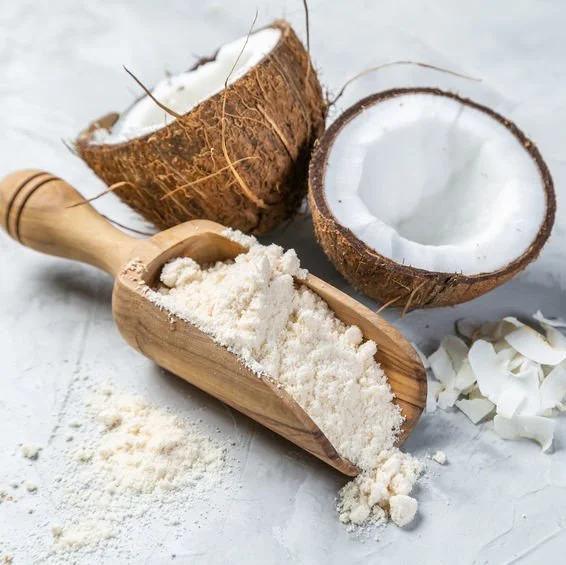 Wooden scoop of coconut flour beside halved coconut — gluten-free, high-fibre baking ingredient.