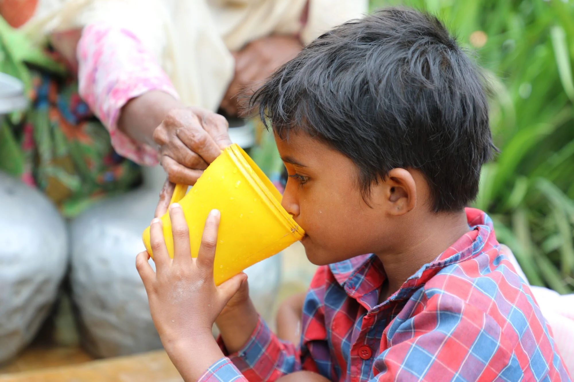 A young boy drinking from a yellow cup outdoors, with an elderly woman pouring a drink into it.