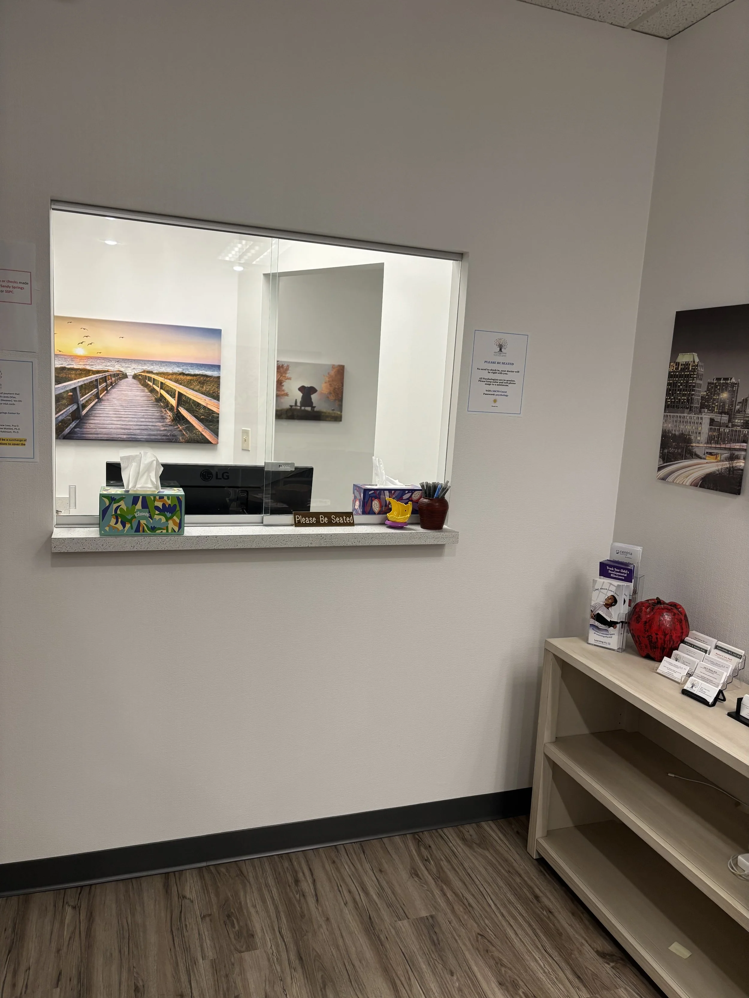 View of a reception window with a computer monitor and a "Please Be Seated" sign, with artwork on the walls and brochures on a wooden shelf nearby.