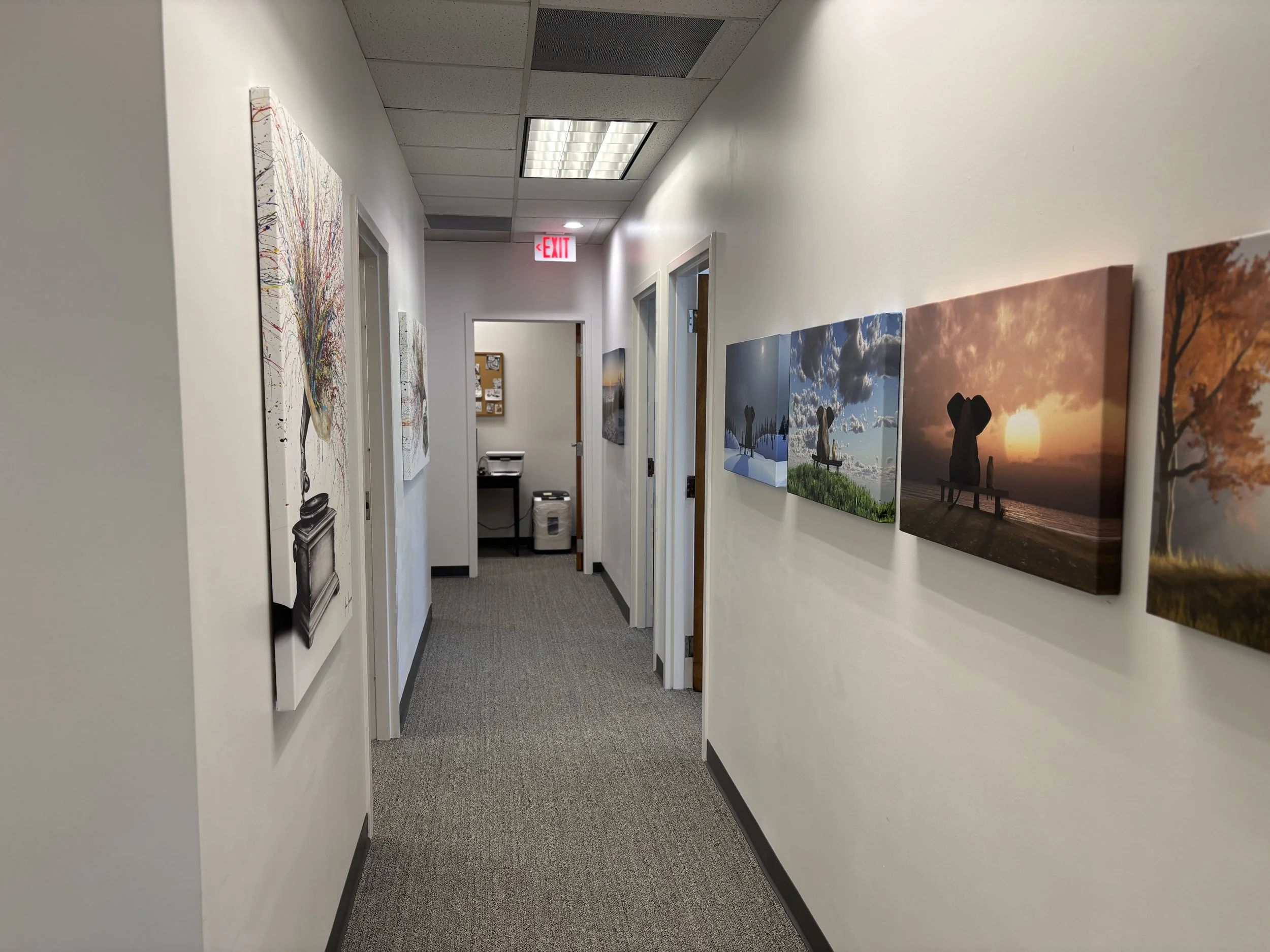 Hallway in an office or gallery with framed artwork on both sides, leading to a small room with a bulletin board and laundry machines, ceiling lights, and an "EXIT" sign.