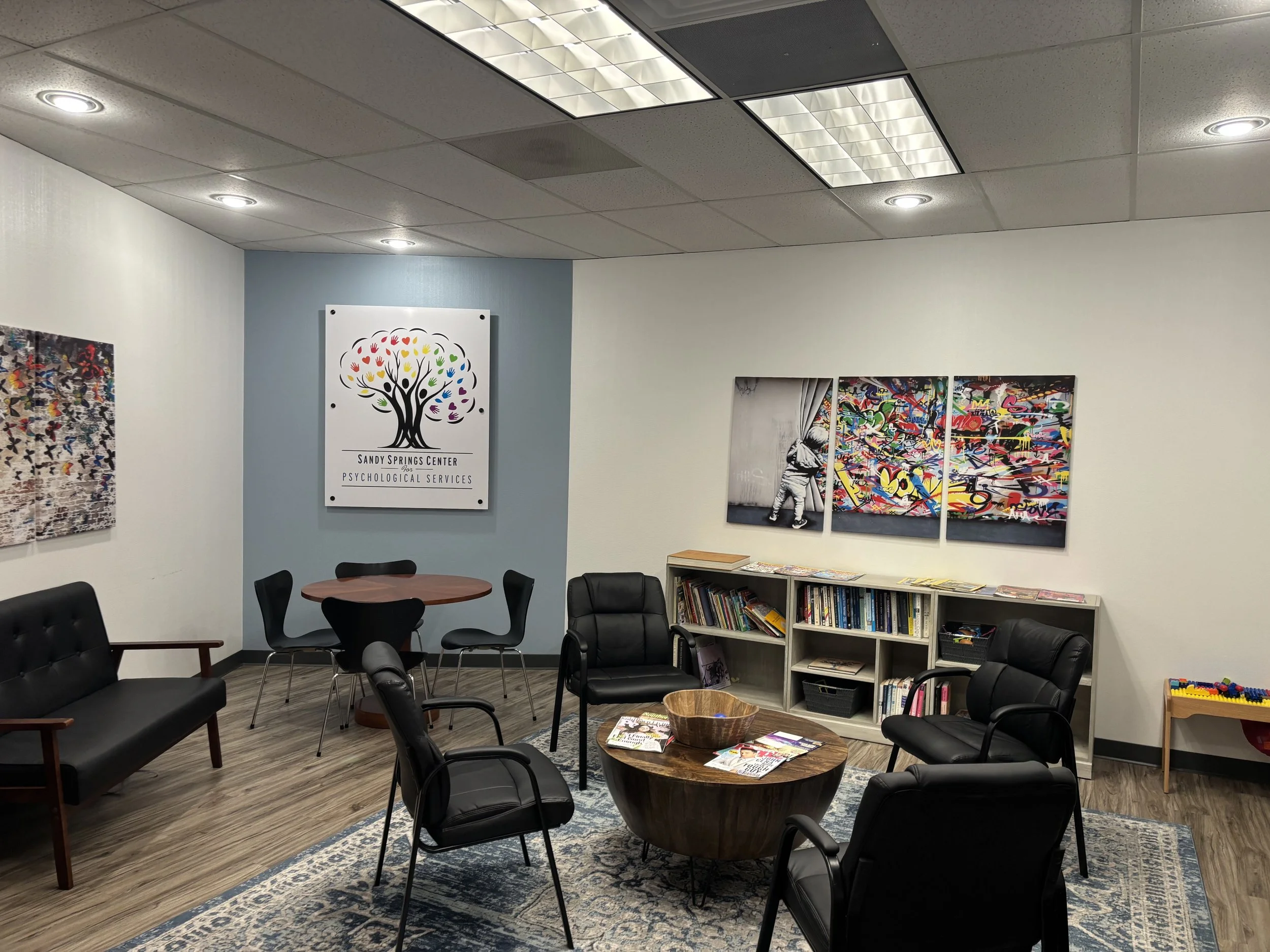 Waiting room with black chairs, a wooden coffee table, bookshelf, colorful artwork, and a sign for Sandy Springs Center Psychological Services on the wall.