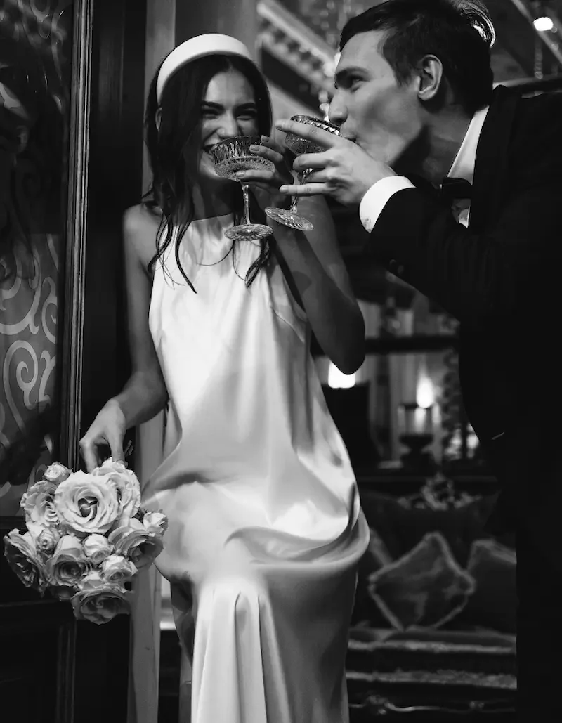 Bride wearing a white slip dress, hairband holding a bouquet of roses smiles with a man in a tuxedo drinks from a glass, sharing a joyful moment.