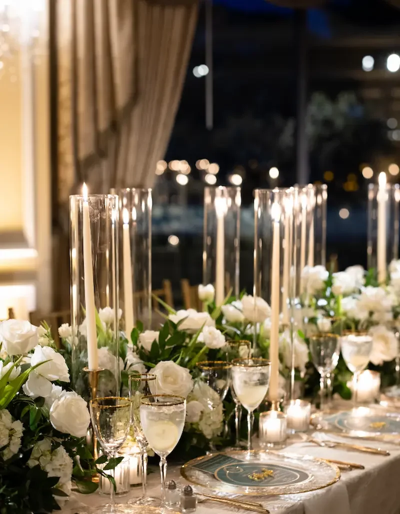 Elegant dining table decorated with white roses, tall candles in glass holders, and fine china, set for an event or celebration.