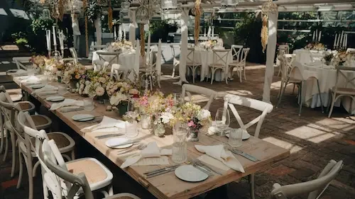 Rustic dinner setup cream coloured chairs, wood tables with pink and white floral arangements down the enter of the table.