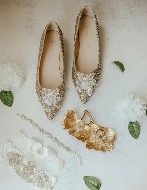 A pair of glittery, pointed-toe wedding shoes with floral embellishments, surrounded by decorative flower jewelry, and wedding accessories on a white surface.