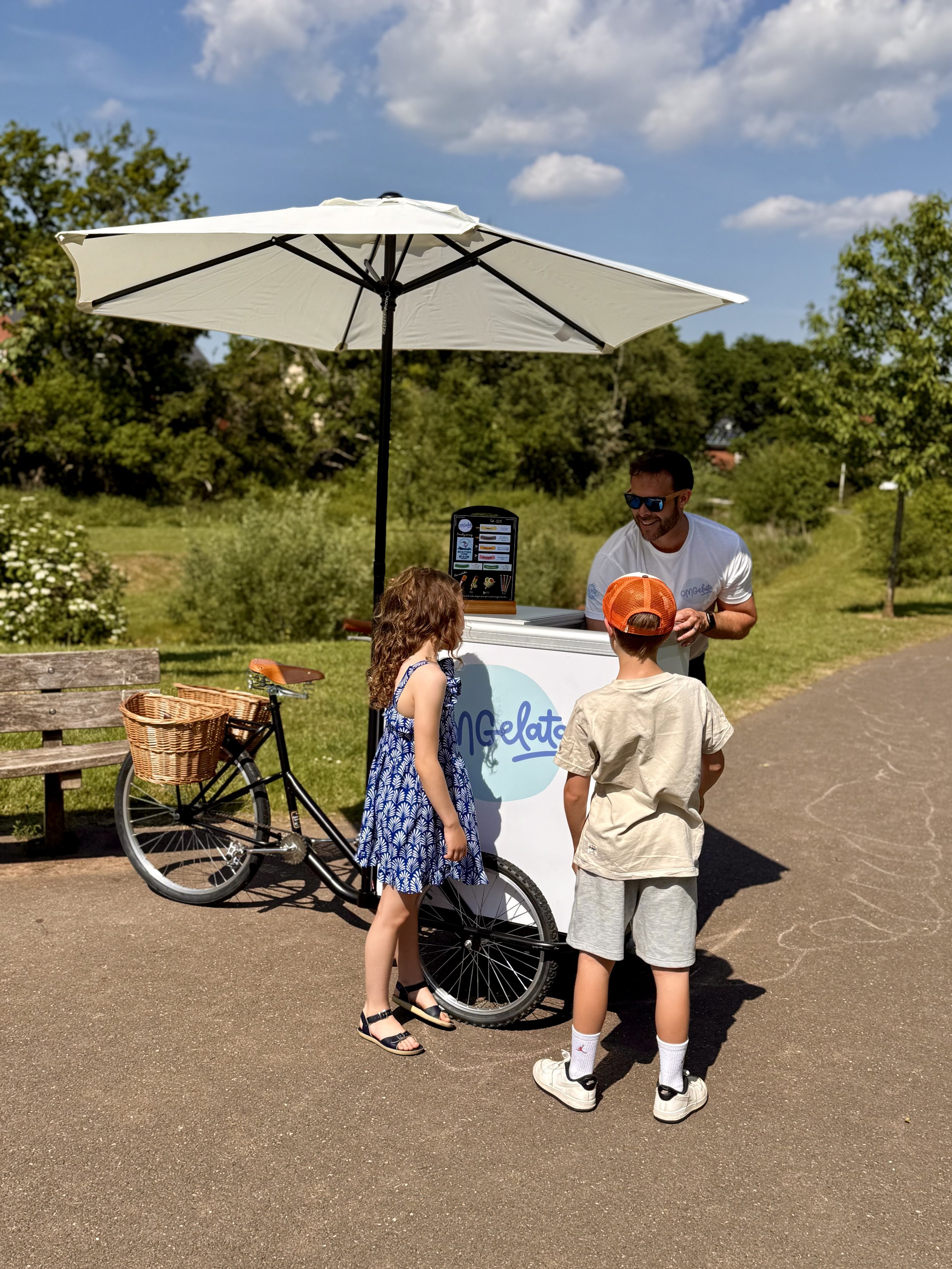 Ice cream vendor with two children ordering ice cream outdoors under a white umbrella, bicycle with basket nearby, sunny day with green trees and a park path in the background.