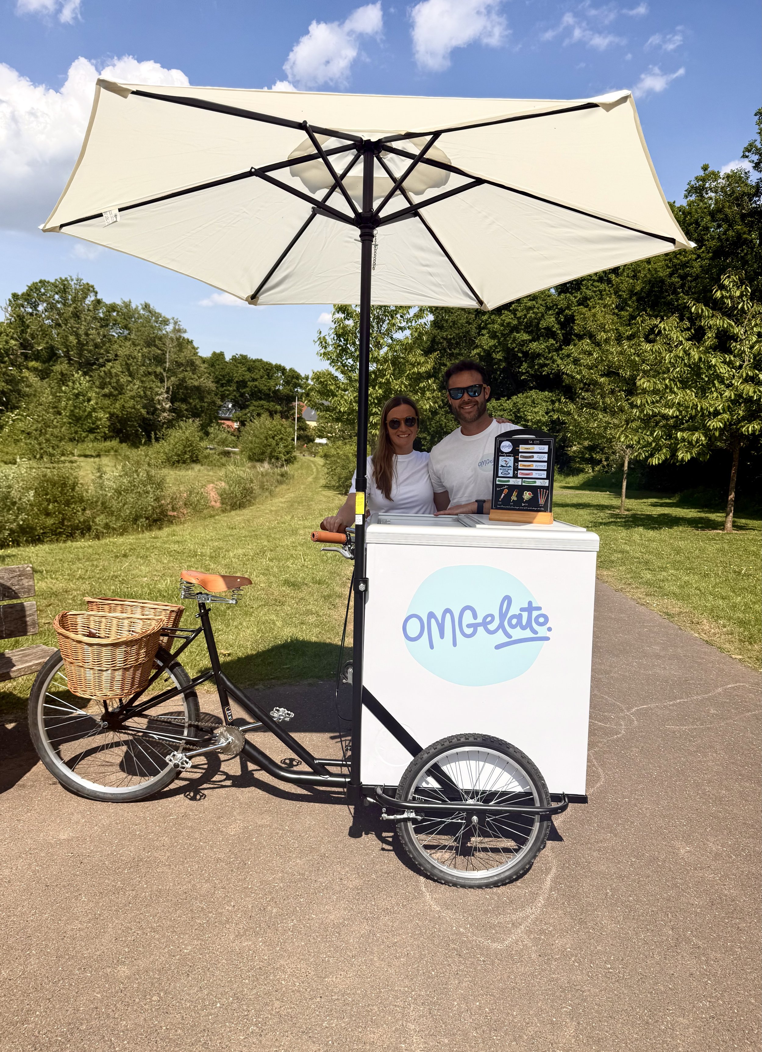 A food cart labeled 'OMGelato' is parked on a park path, shaded by a large white umbrella, with two smiling people standing behind it. The fruit bike has a basket on the front. Trees and blue sky are in the background.