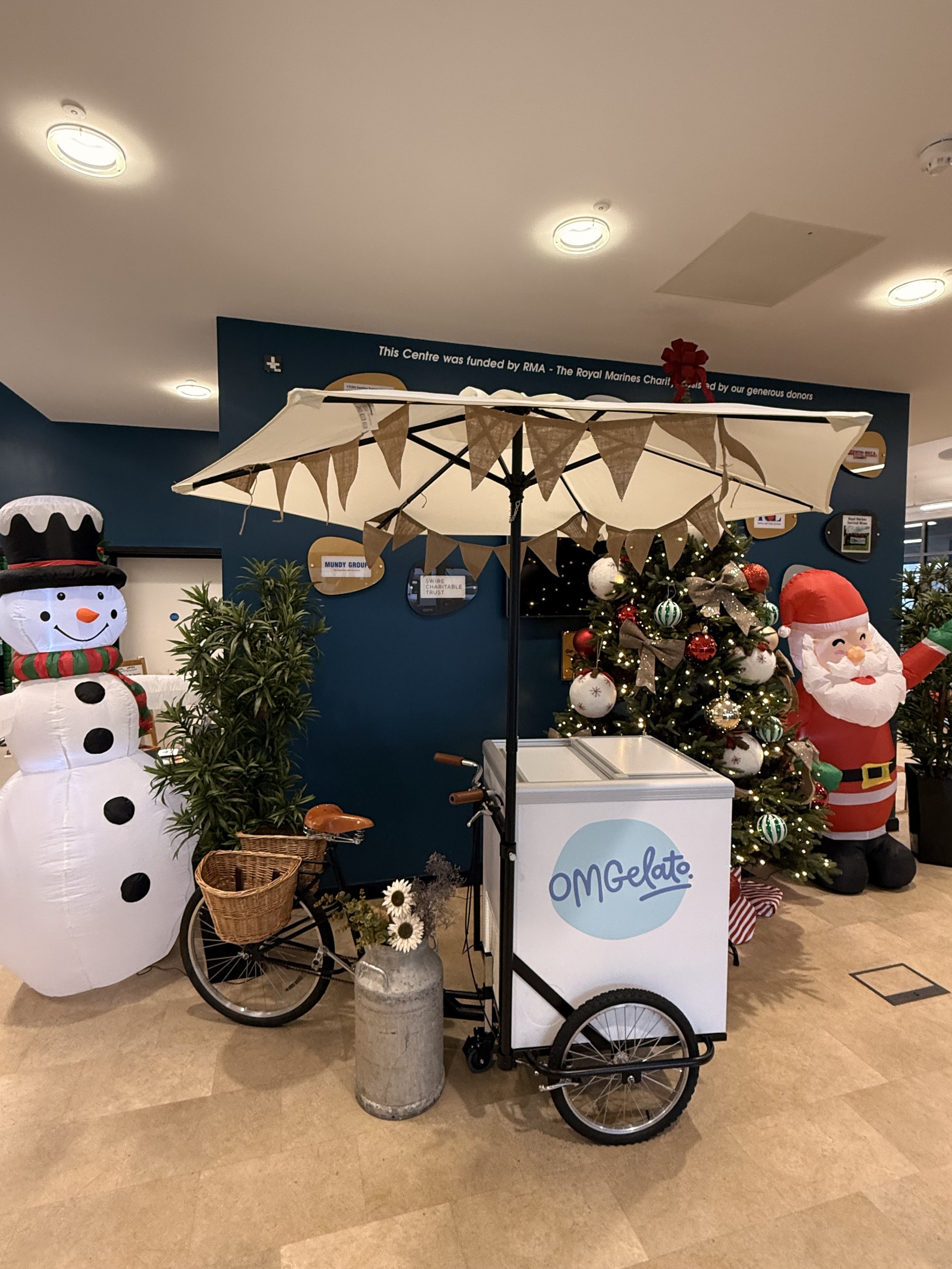 A Christmas-themed display featuring a snowman, a decorated Christmas tree, a Santa Claus figure, an OM Gelato cart with an umbrella, and some plants in the background.