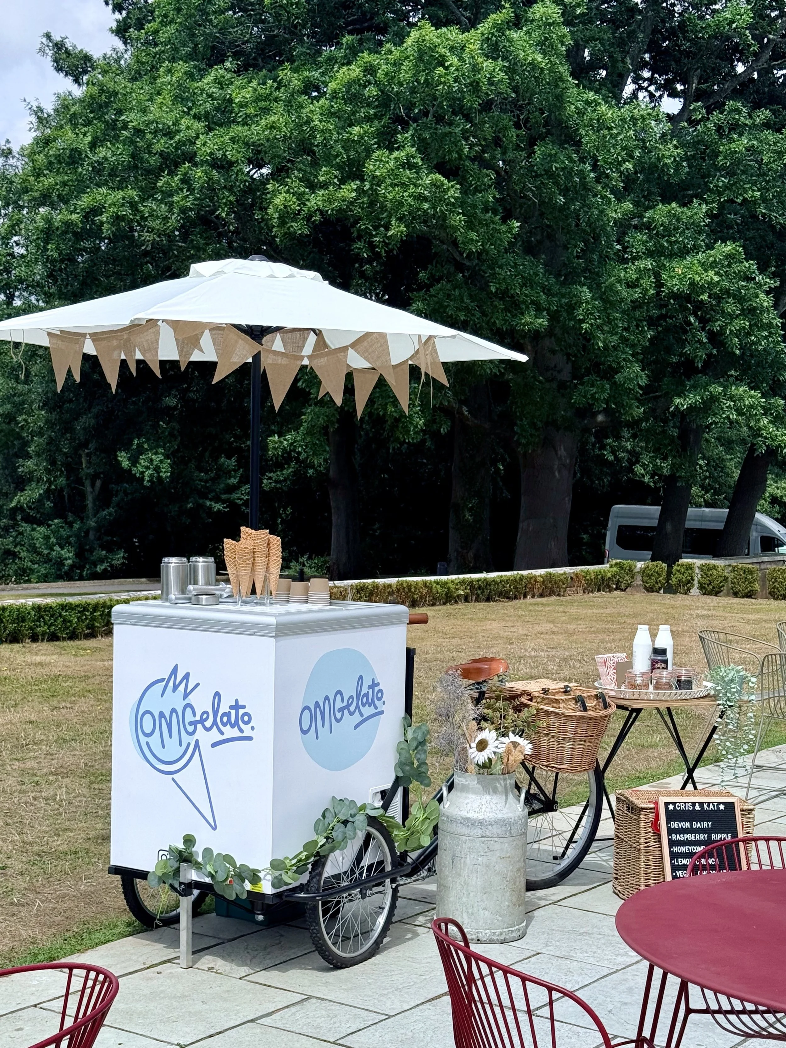 Ice cream cart with a white umbrella, outdoor seating with red chairs, various food and drink items, and a large tree in the background.