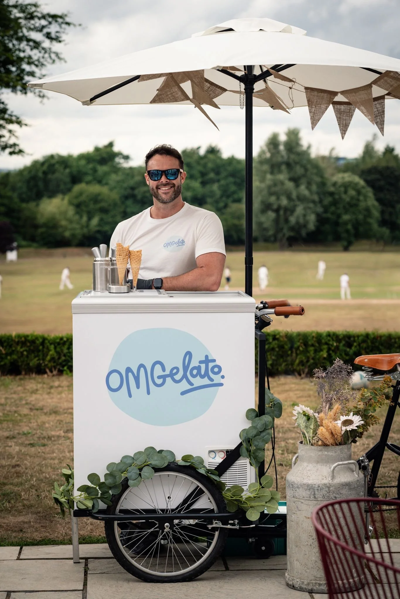 A man with sunglasses and a white t-shirt stands behind an ice cream cart labeled 'OMGelato'. The cart has a large umbrella, and there are ice cream cones on display. The man is smiling, and in the background, people are playing cricket on a field with trees.