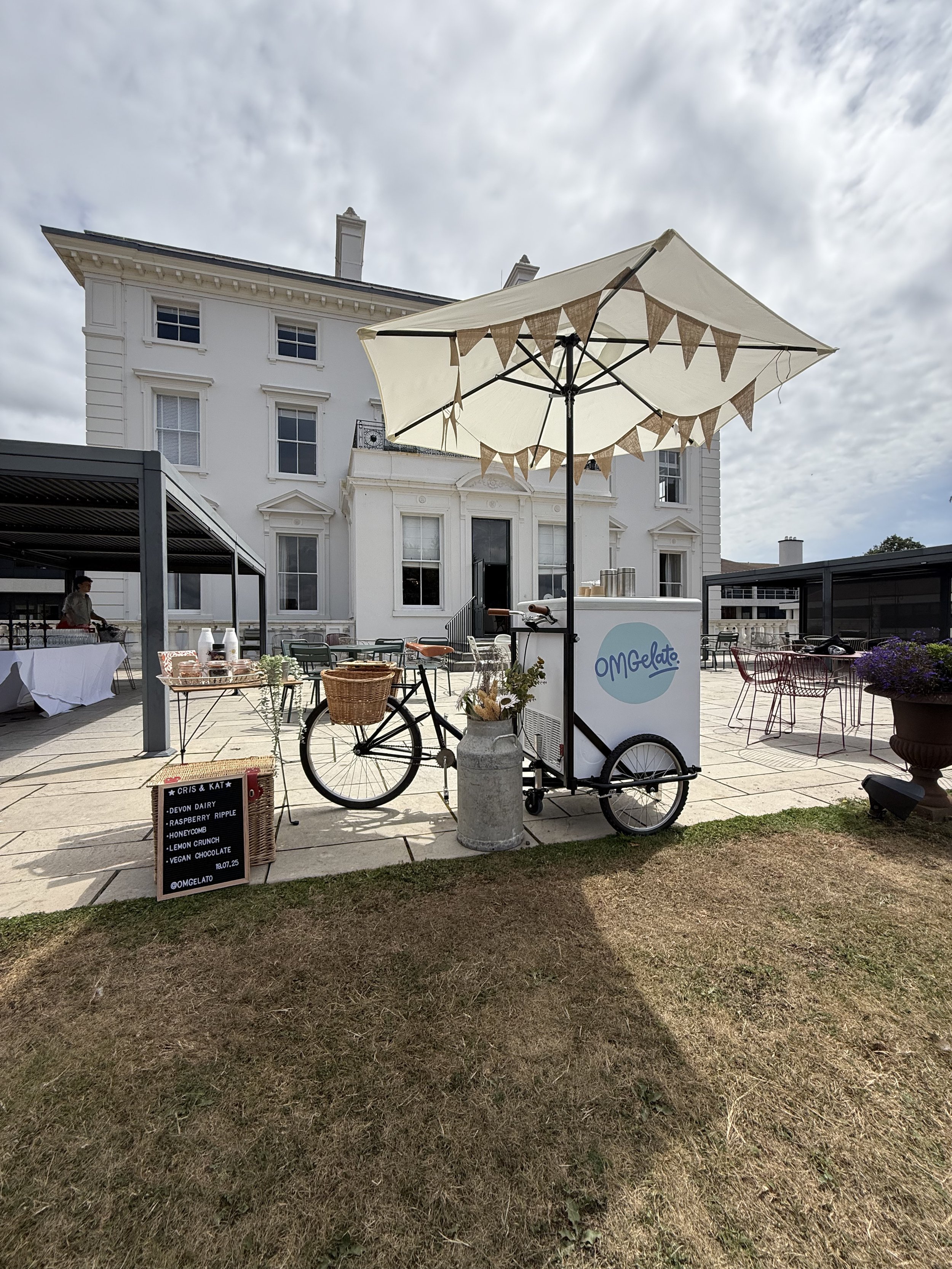 Ice cream cart with a beige umbrella outside with a house in the background.