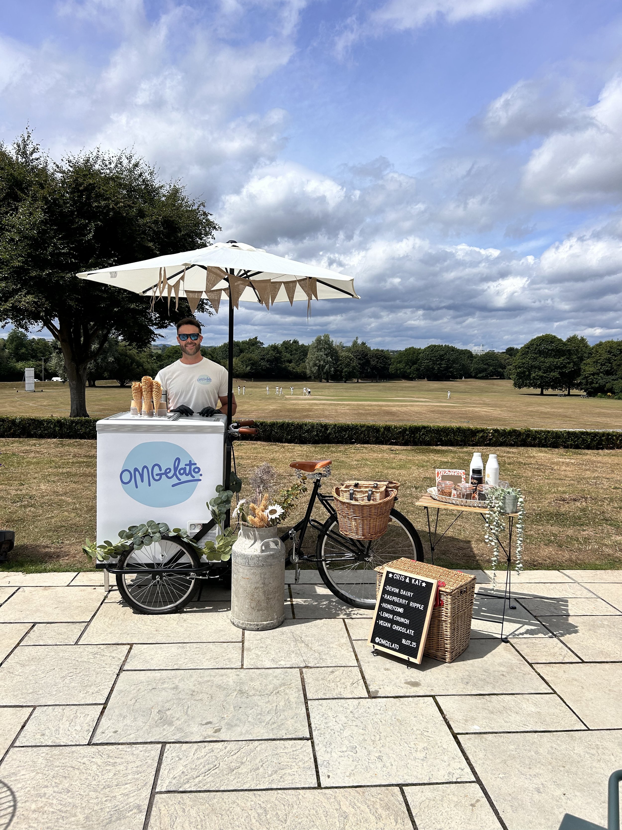 A man with sunglasses stands behind a mobile ice cream cart under a white umbrella in an outdoor park, with tables, a bicycle, and a chalkboard sign nearby.