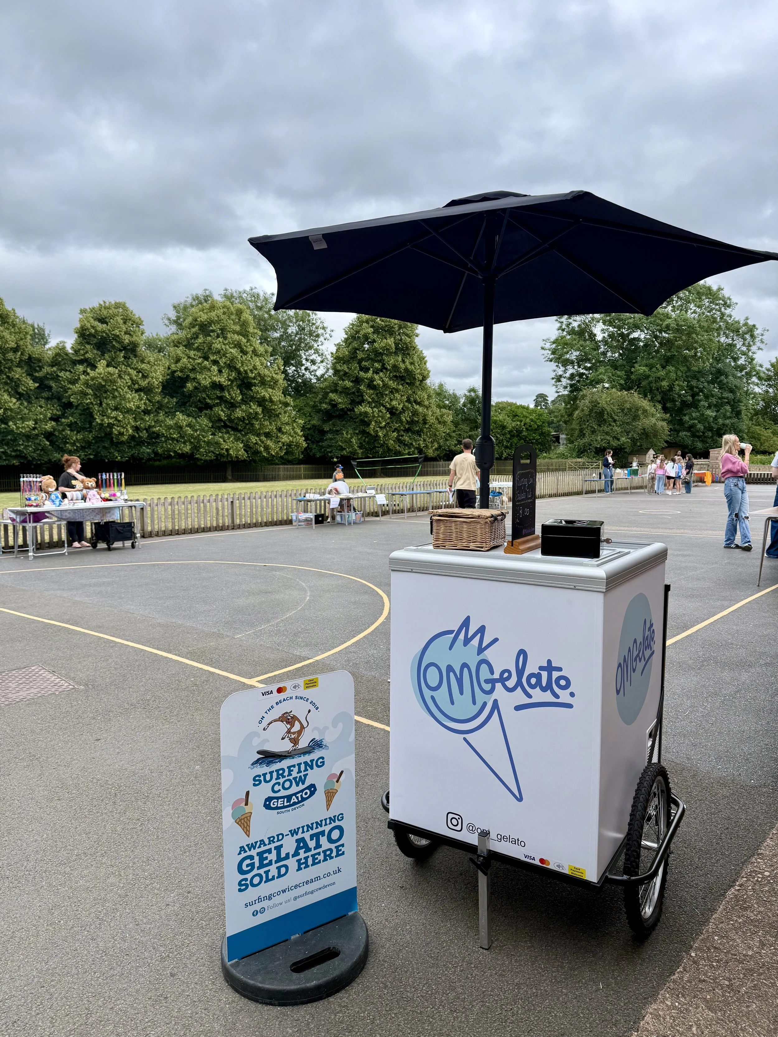 Ice cream cart with black umbrella, advertising 'OMG Elato' ice cream, positioned on an outdoor court, with people shopping at nearby stalls and trees in the background.