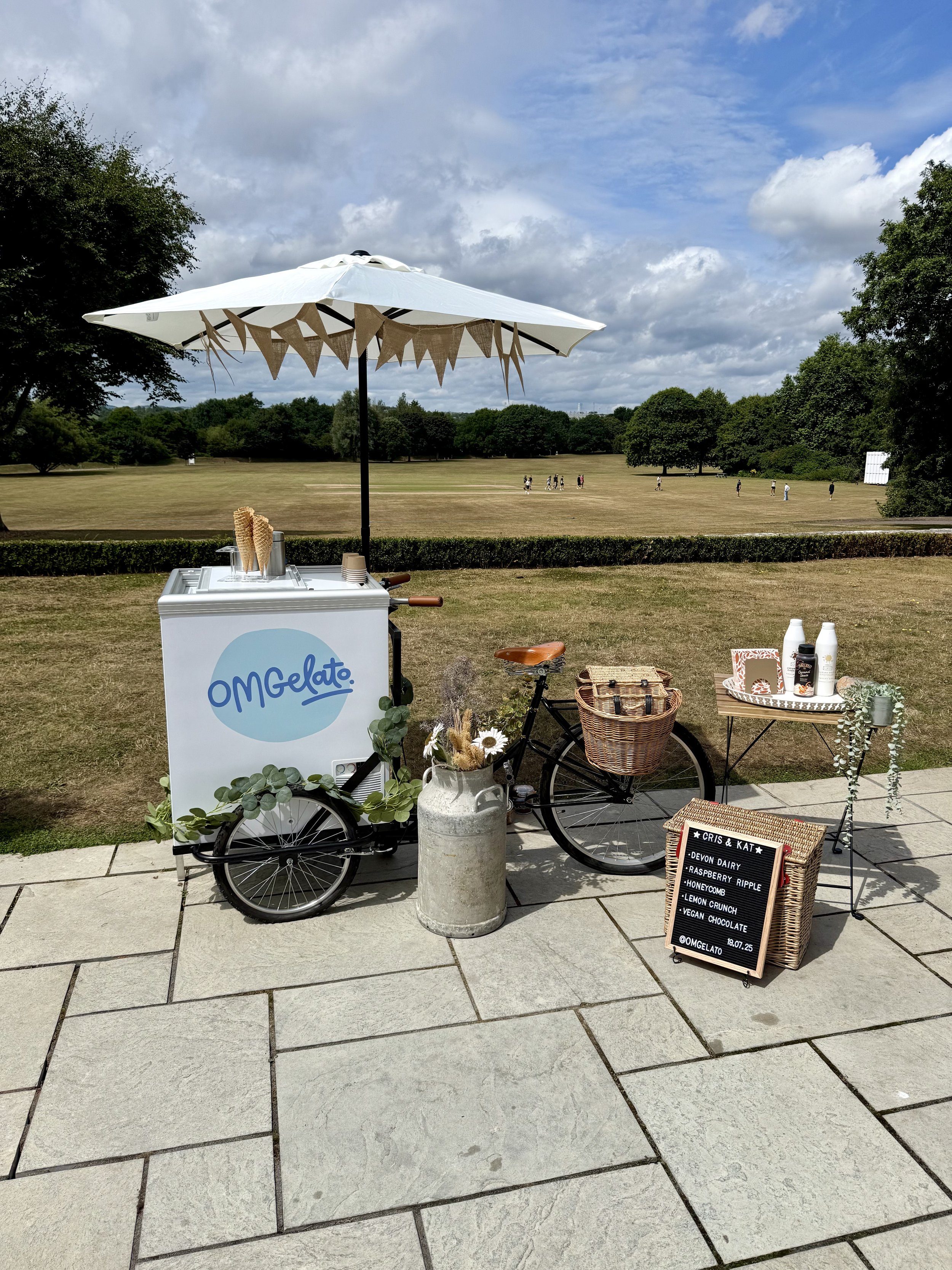 Ice cream cart with an umbrella and decorative bunting, set up outdoors on a paved area next to a grassy park with trees and people in the background. The cart displays the OM Gelato logo, and there is a small table with bottles and a sign listing fl