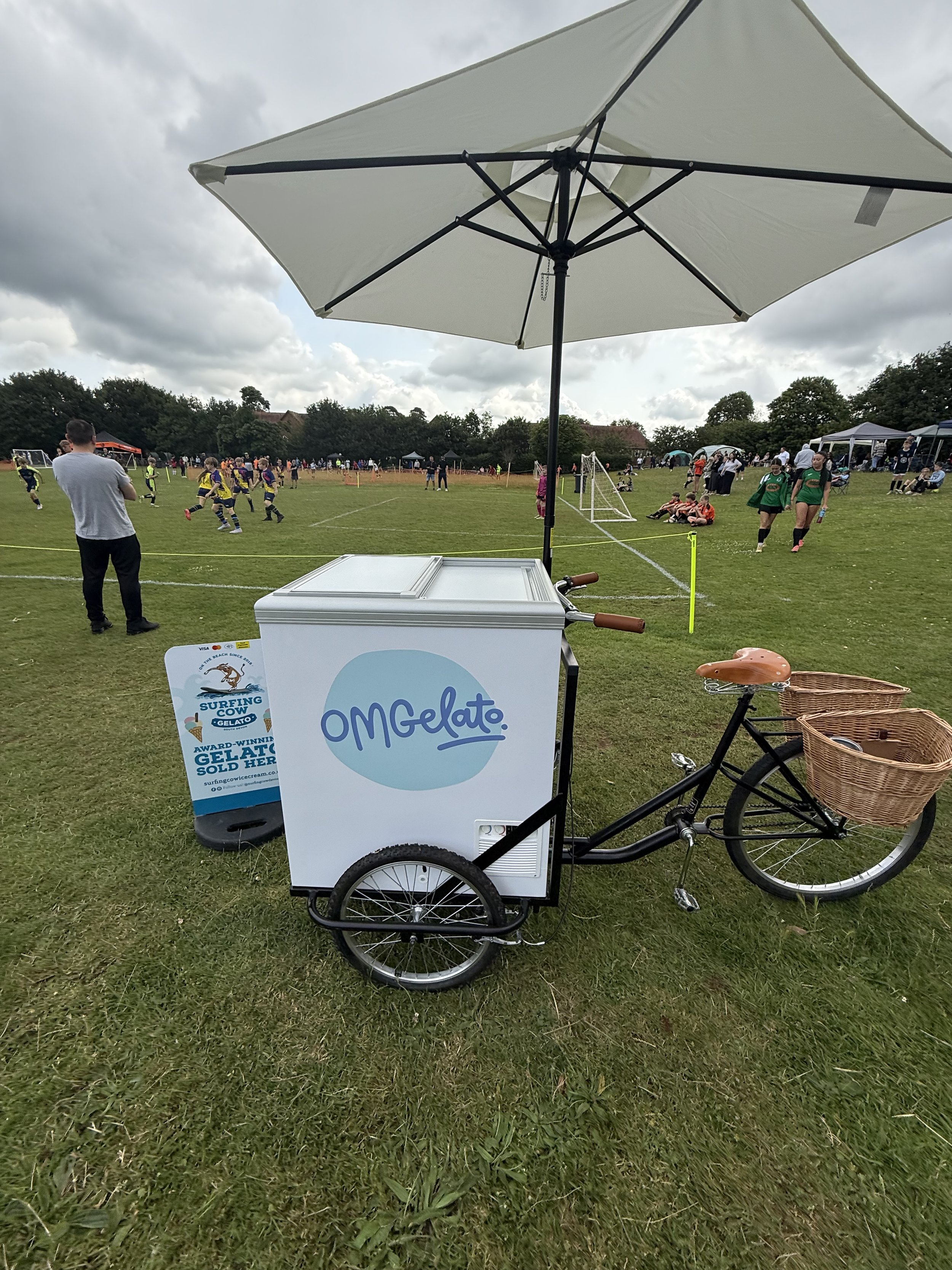 A gelato and ice cream tricycle with a large umbrella on a grassy field during a sports event.