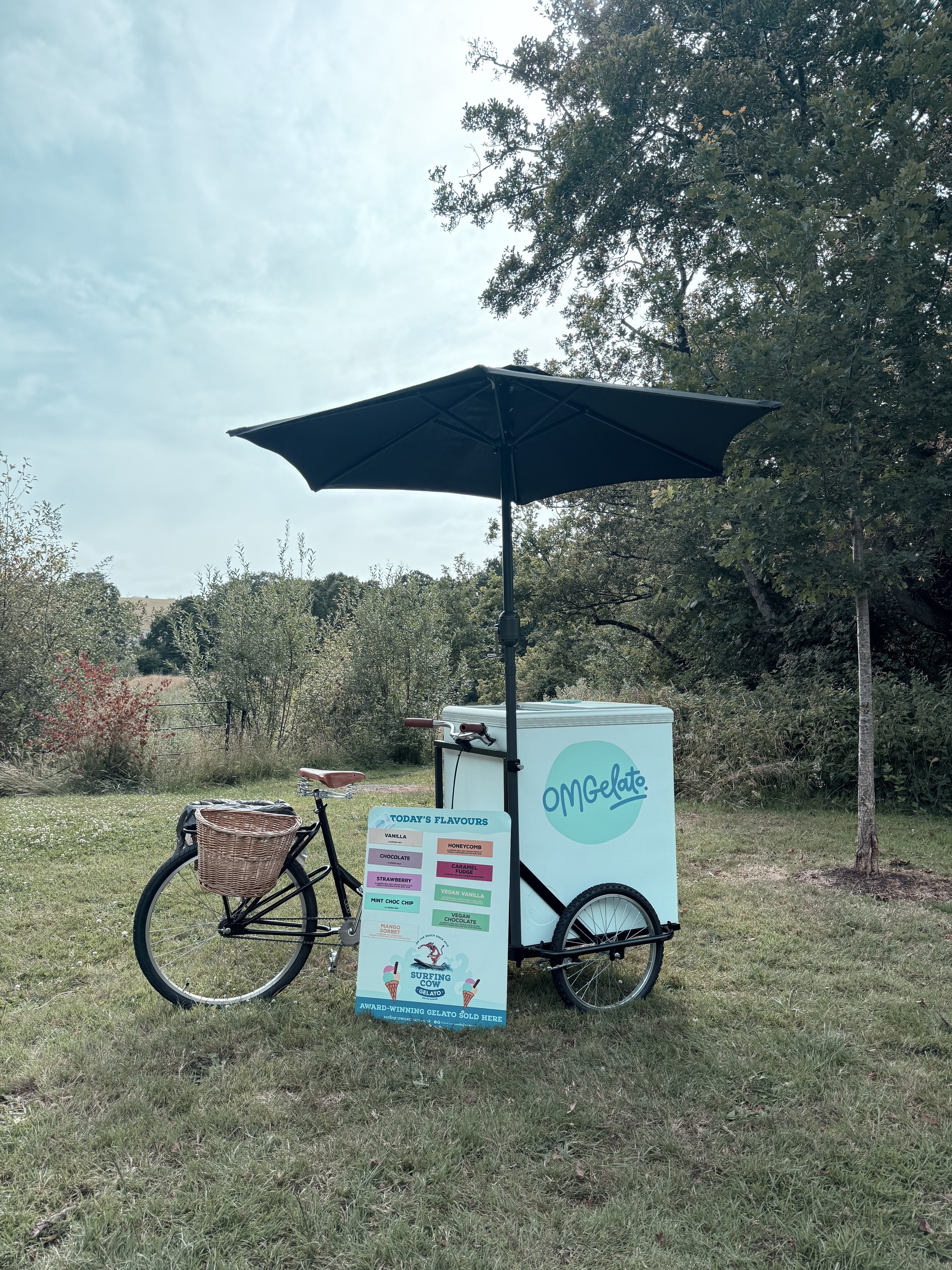 A bicycle with a wicker basket parked next to a gelato cart under a large black umbrella outdoors. The gelato cart has a sign listing flavors and the word 'Omg!! Gelato' on the side.