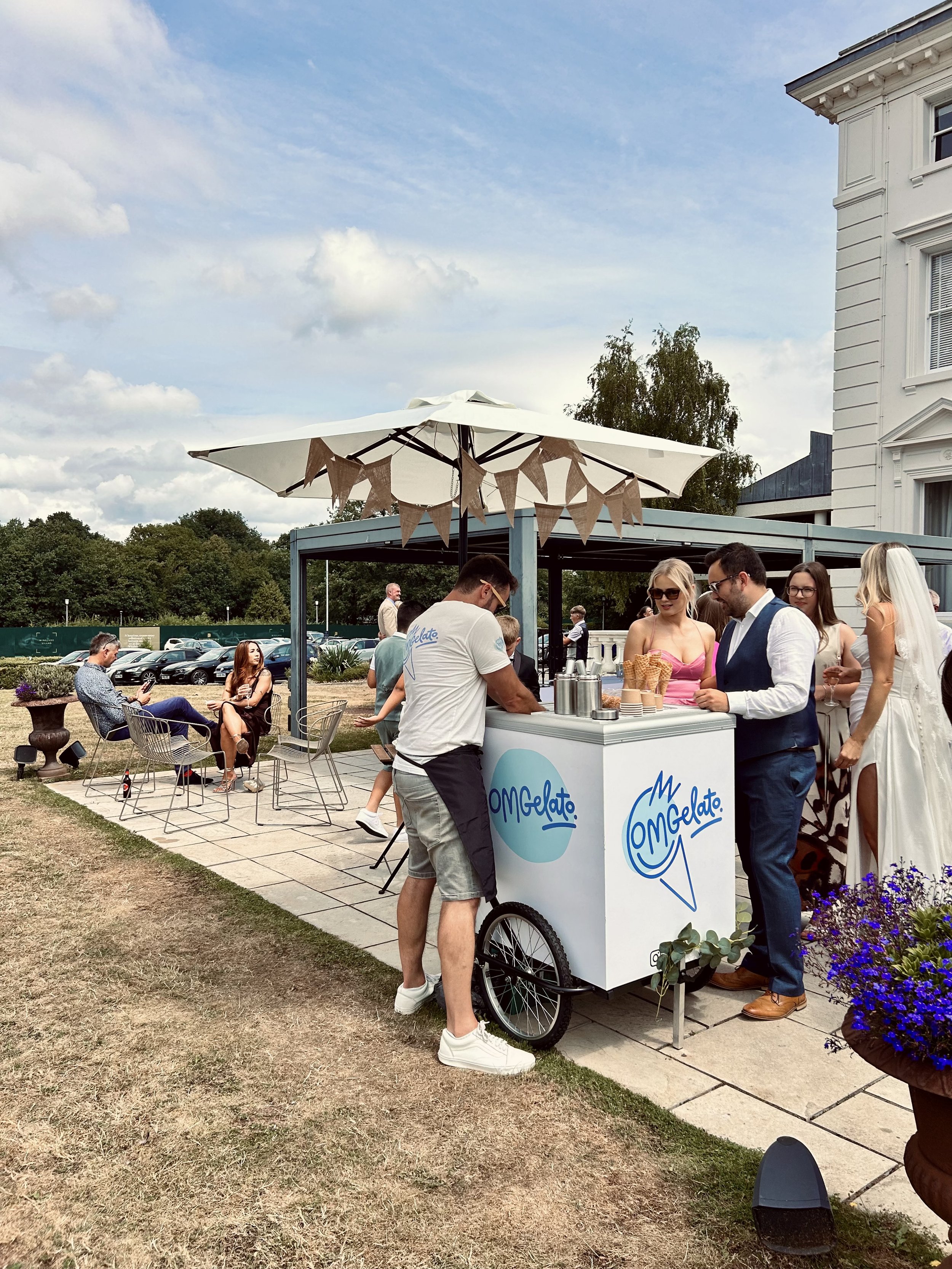 People gathered at an outdoor ice cream cart with a white umbrella, with a sign that says 'M Gelato,' on a sunny day. Some people are sitting at tables, while others order ice cream at the cart. There is a white building with decorative trim on the r