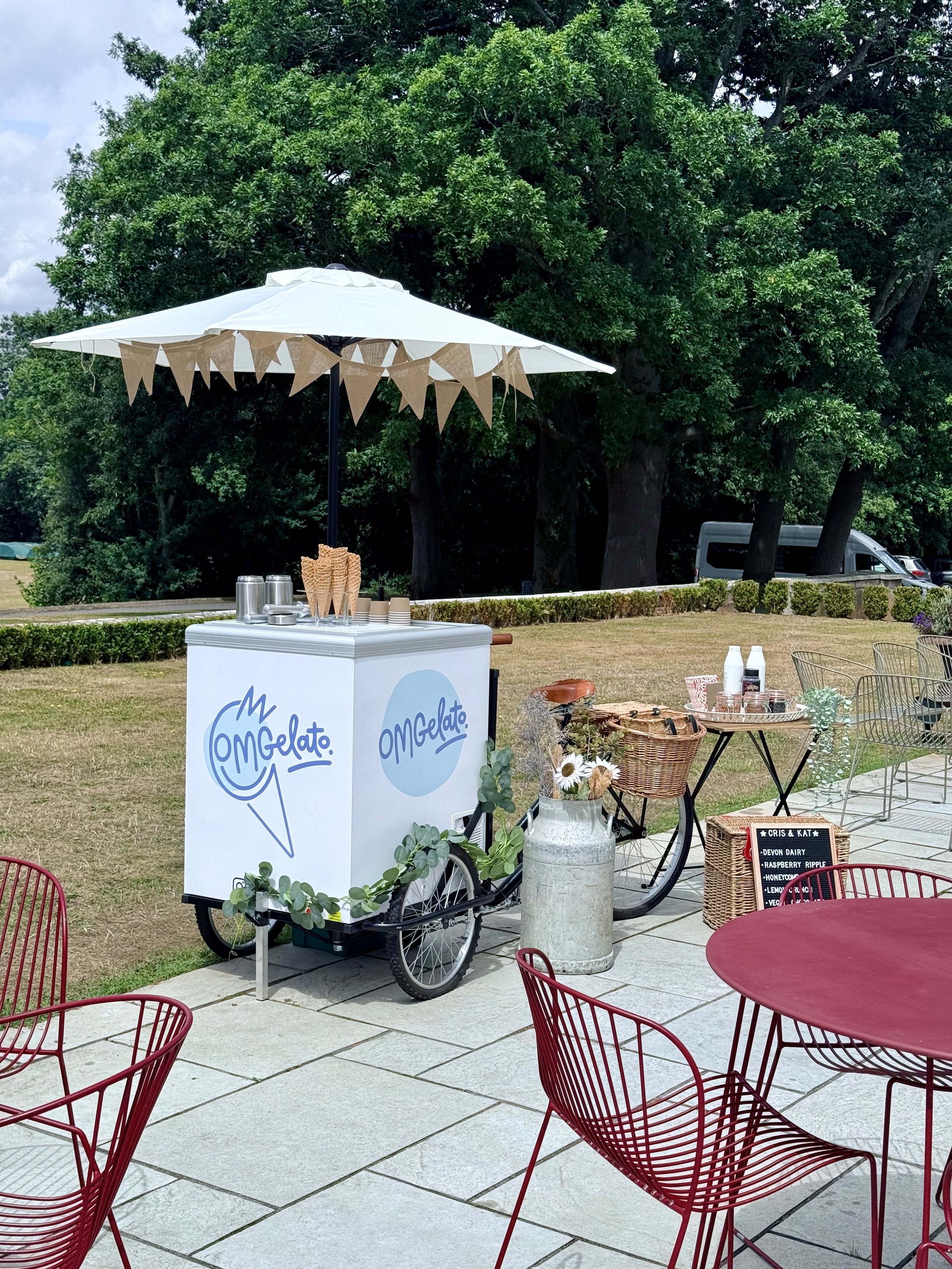 An ice cream cart with 'Omg Gelato' written on it, set up outdoors under a large white umbrella decorated with bunting. The cart has ice cream cones, cups, and toppings, with tables and chairs nearby, and a blackboard sign listing flavors.