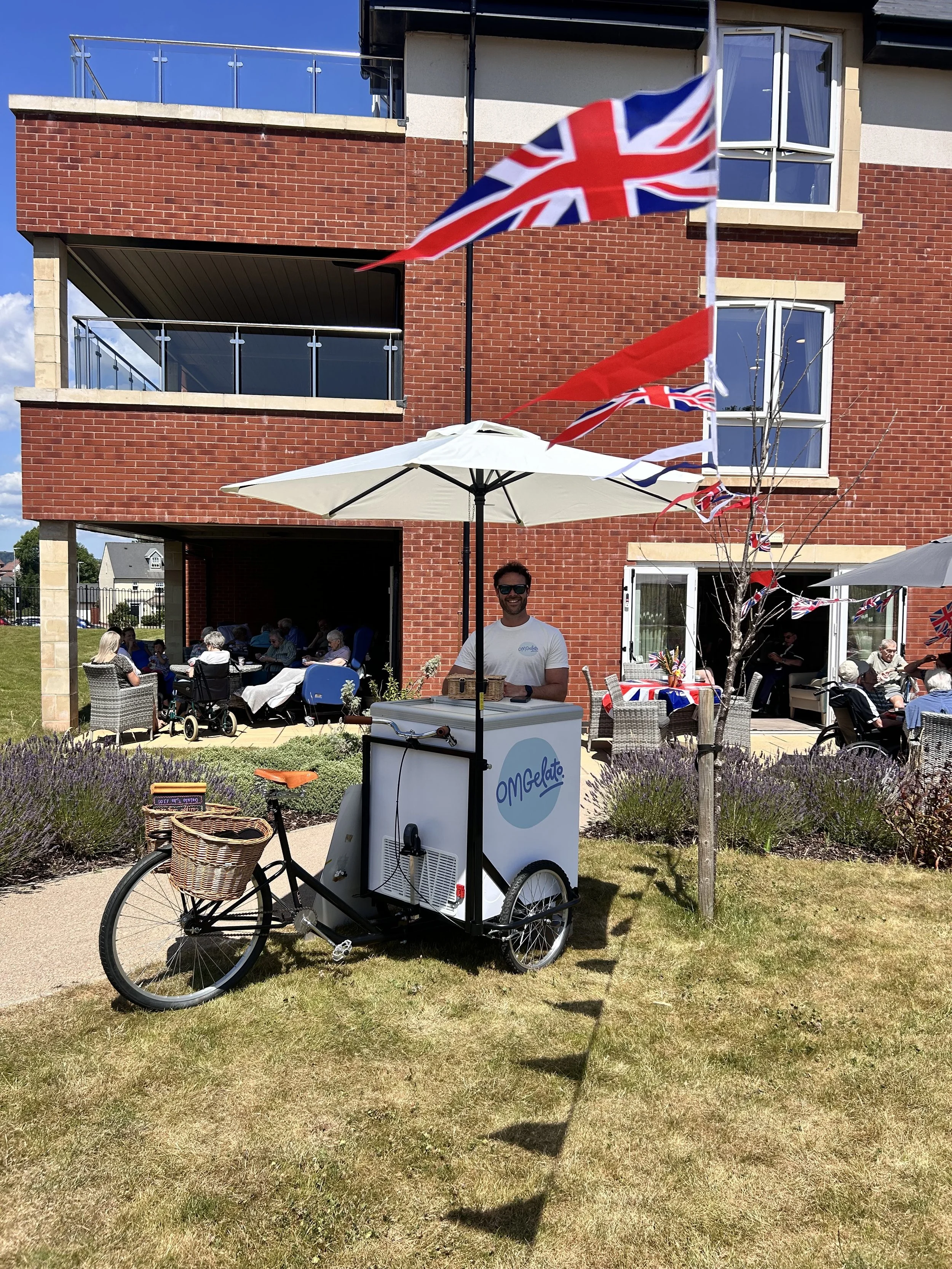 A man standing next to a mobile ice cream or gelato cart on a bike with a basket, in front of a brick apartment building with patio seating, flags, and people enjoying the outdoor gathering.